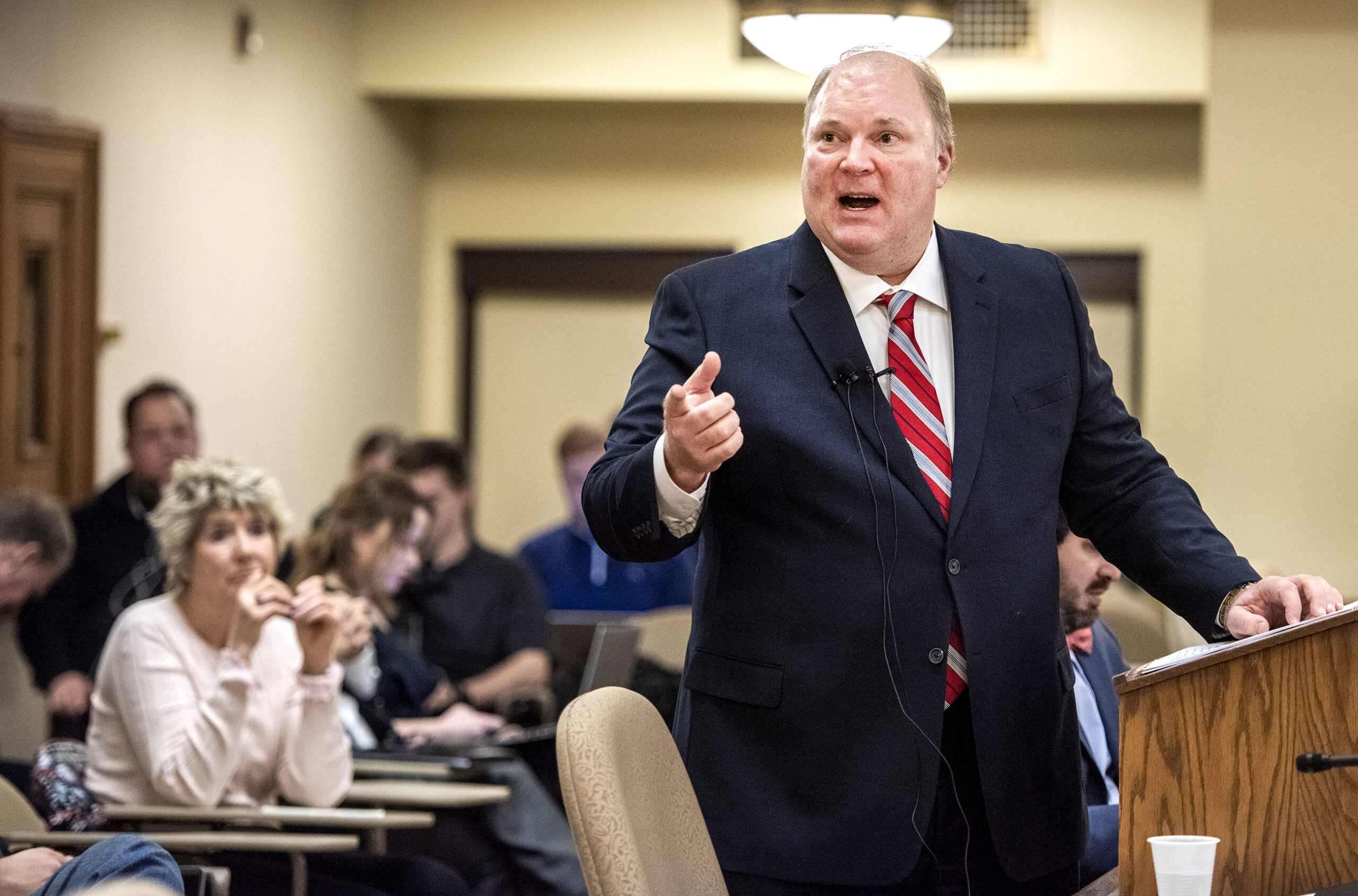 Michael Gableman stands at a podium and points around the room as he speaks. Attendees are seated behind him.