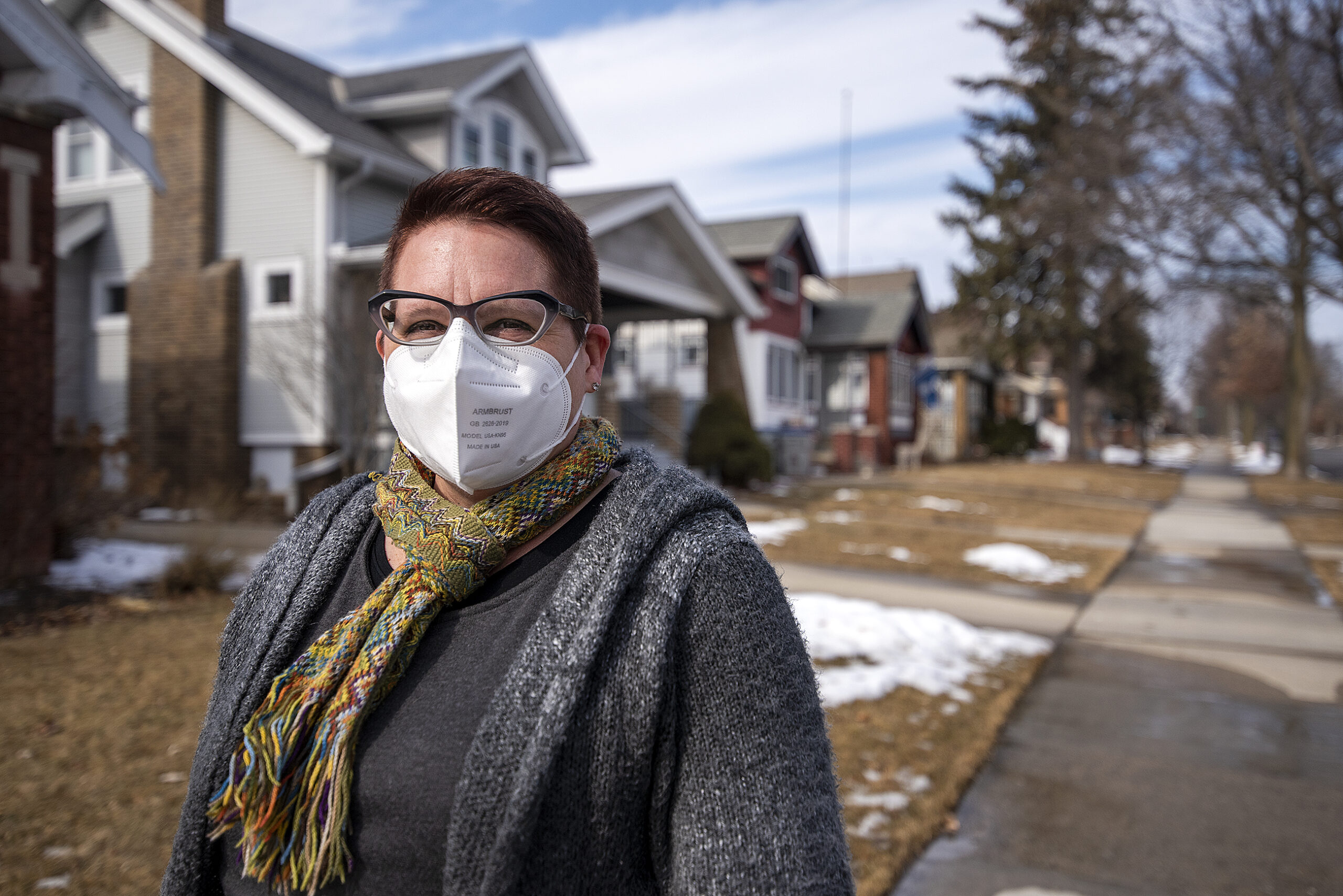 A woman stands near a row of homes outside wearing a white face mask.