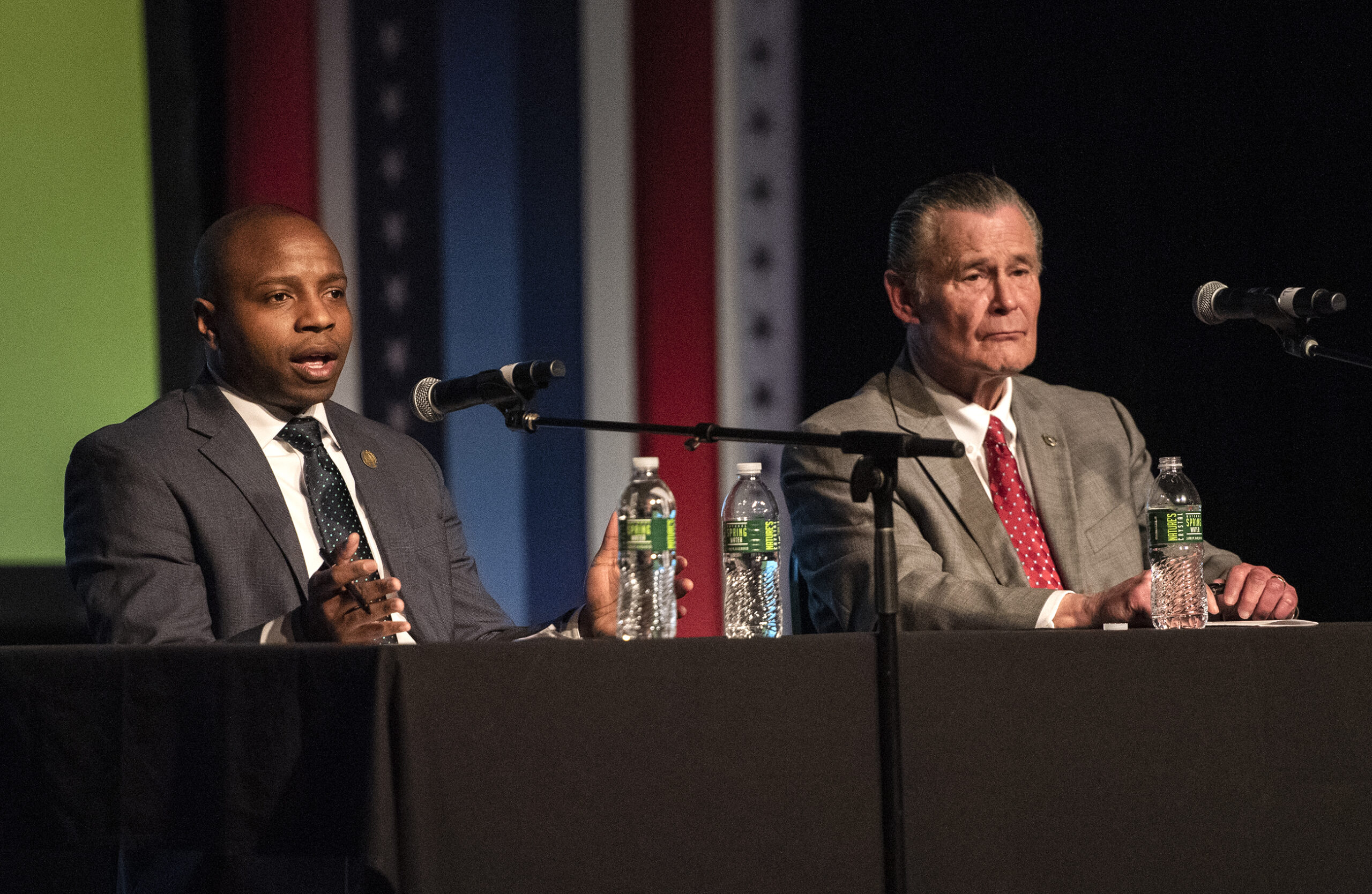 Two candidates sit on stage in front of microphones.