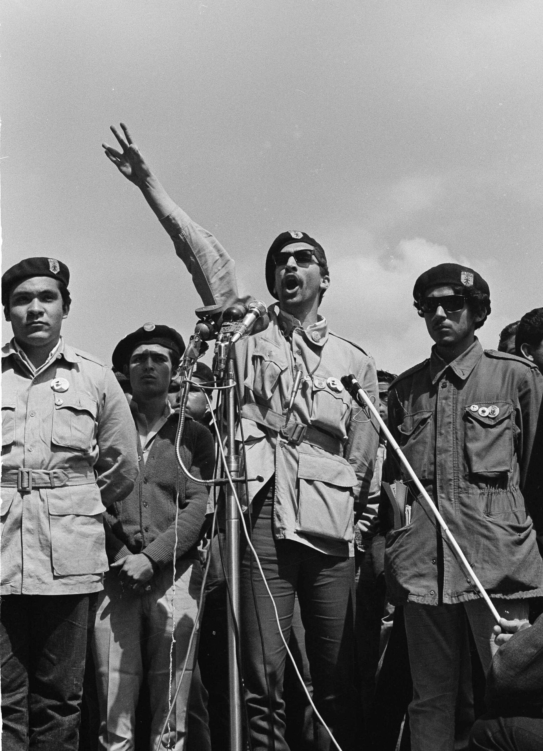 A co-founder of the Brown Berets speaks at a microphone with one arm raised
