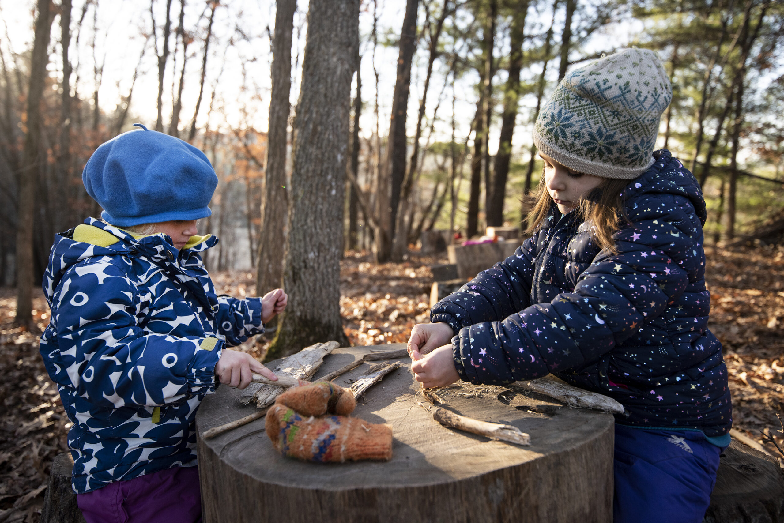 Two girls in winter coats and hats play at a tree stump