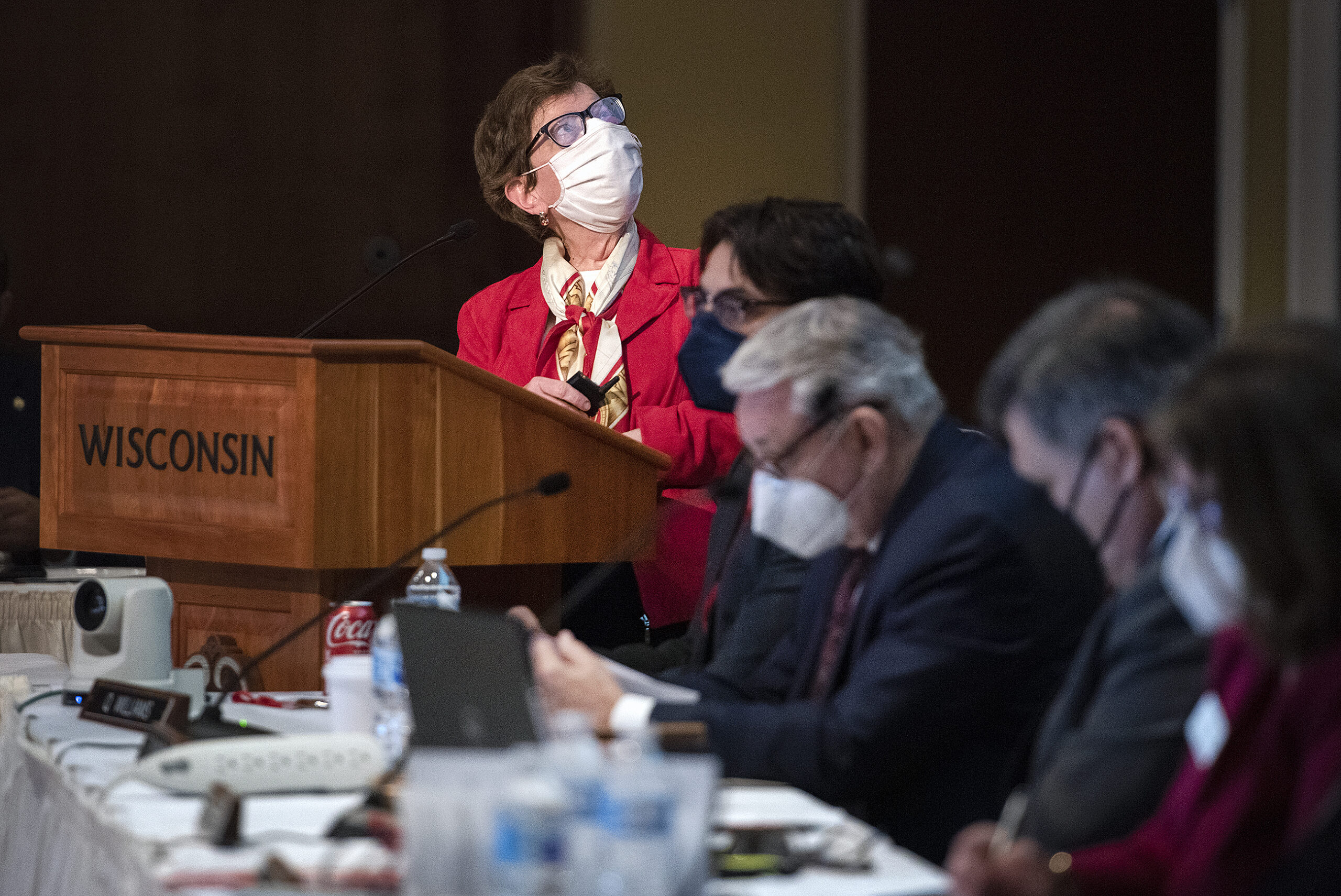 Rebecca Blank stands at a podium while speaking. She wears a white face mask and a red jacket.