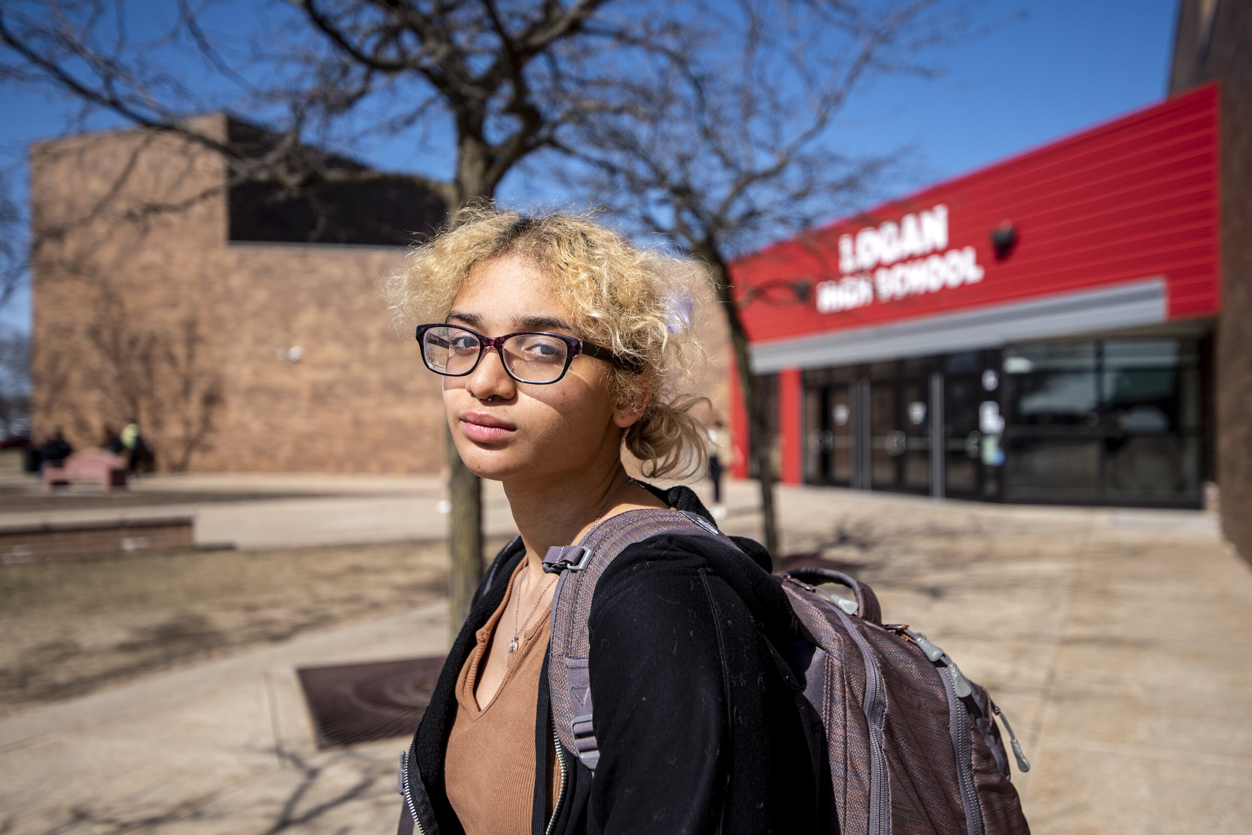 A student wears her backpack as she stands outside of her school on a sunny day.