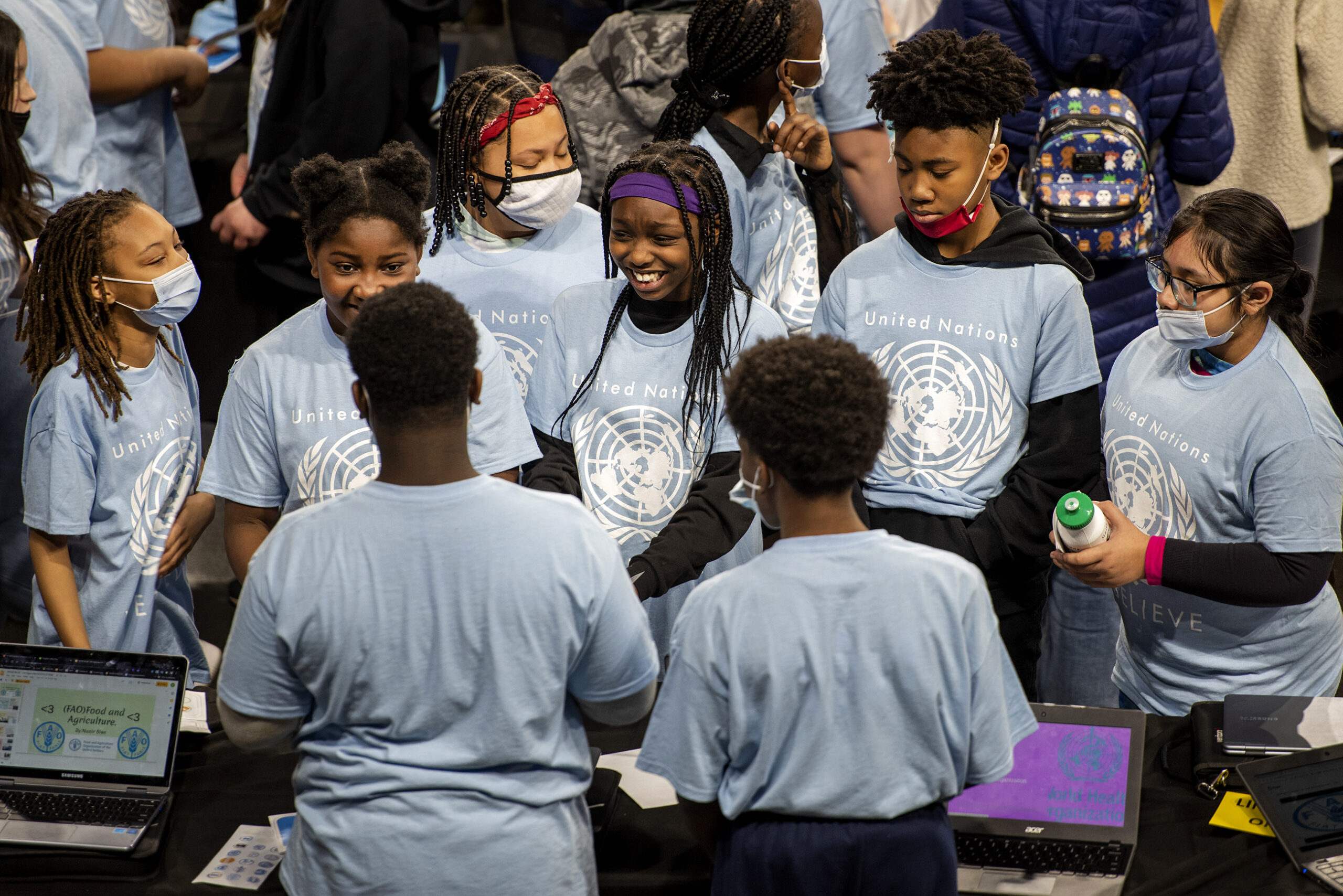 Students in matching t-shirts gather around a table to hear a presentation.