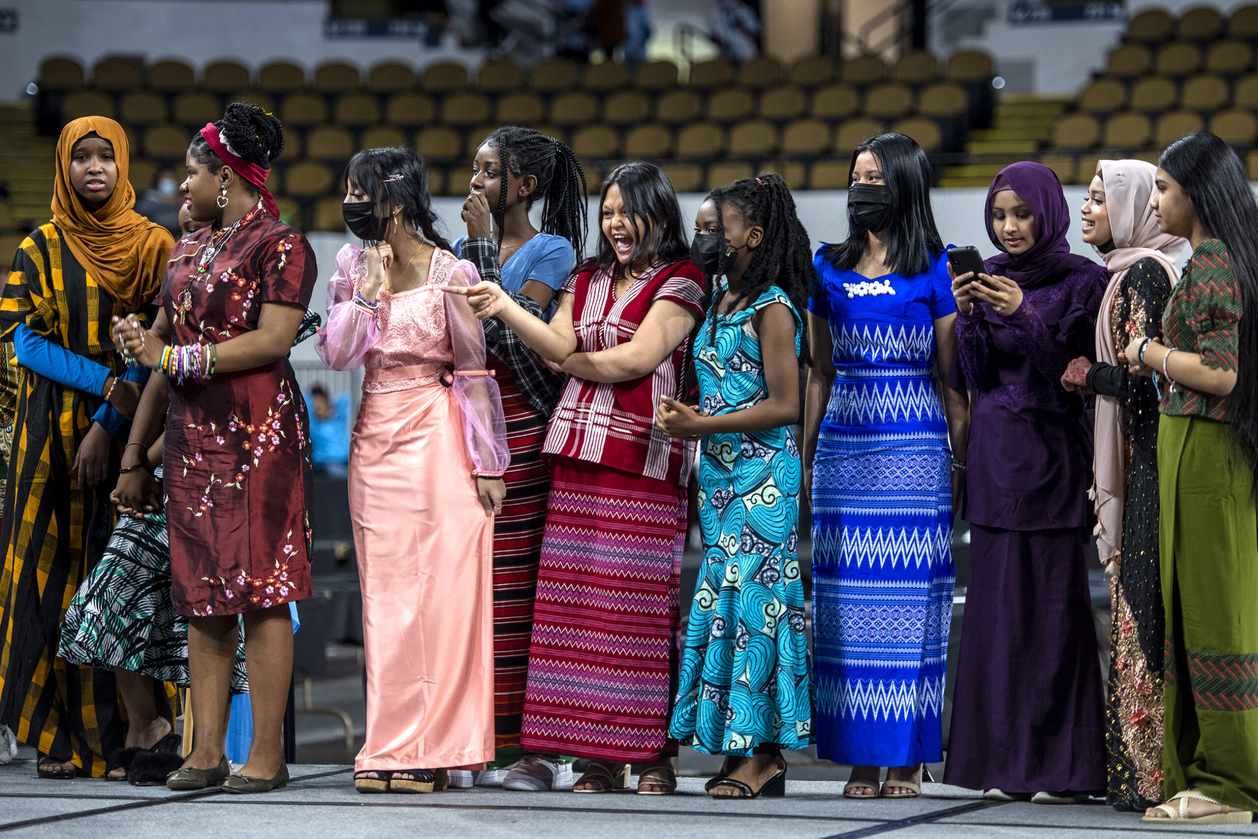 Students in colorful clothing representing different countries watch as their fellow classmates walk on stage and explain the culture behind their clothing to a crowd.