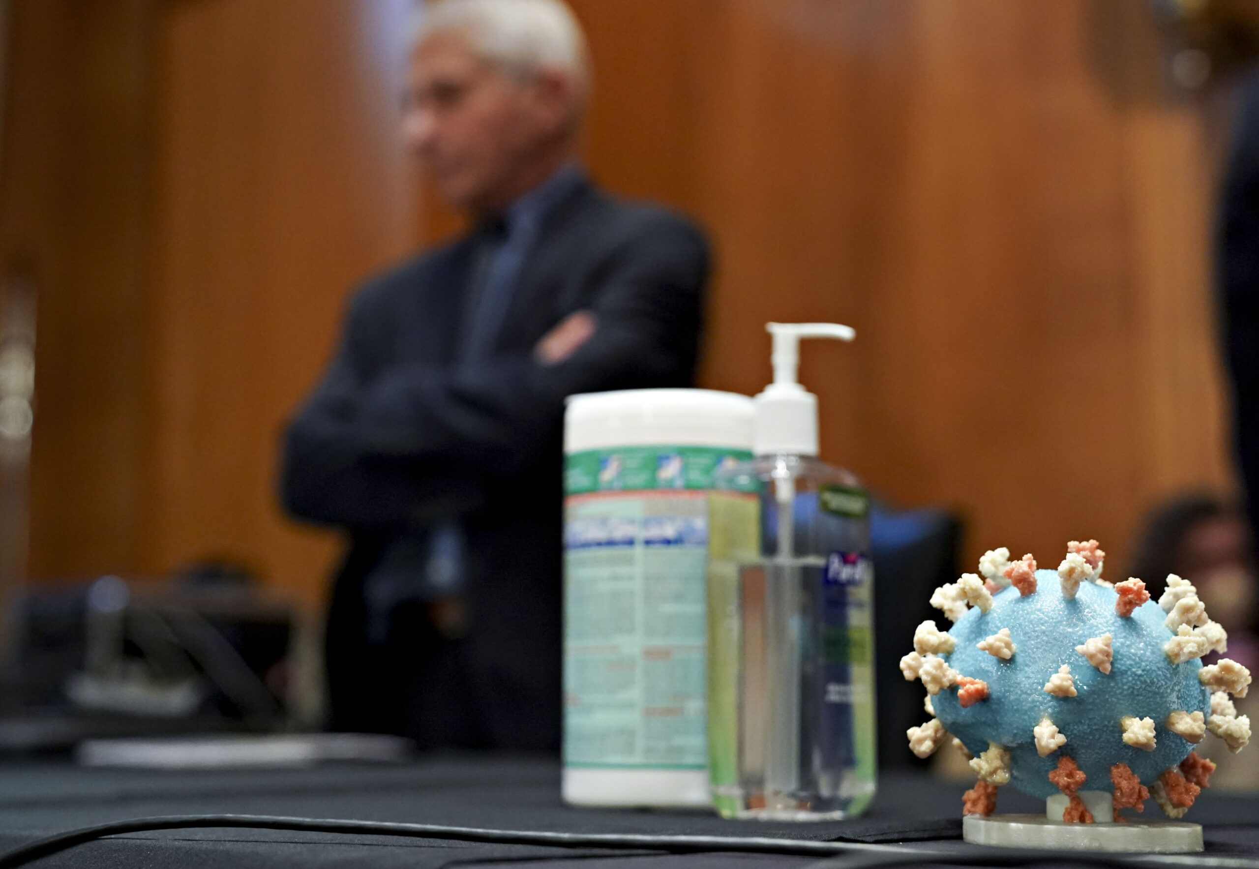 Hand sanitizer and coronavirus model on a table with Fauci in background