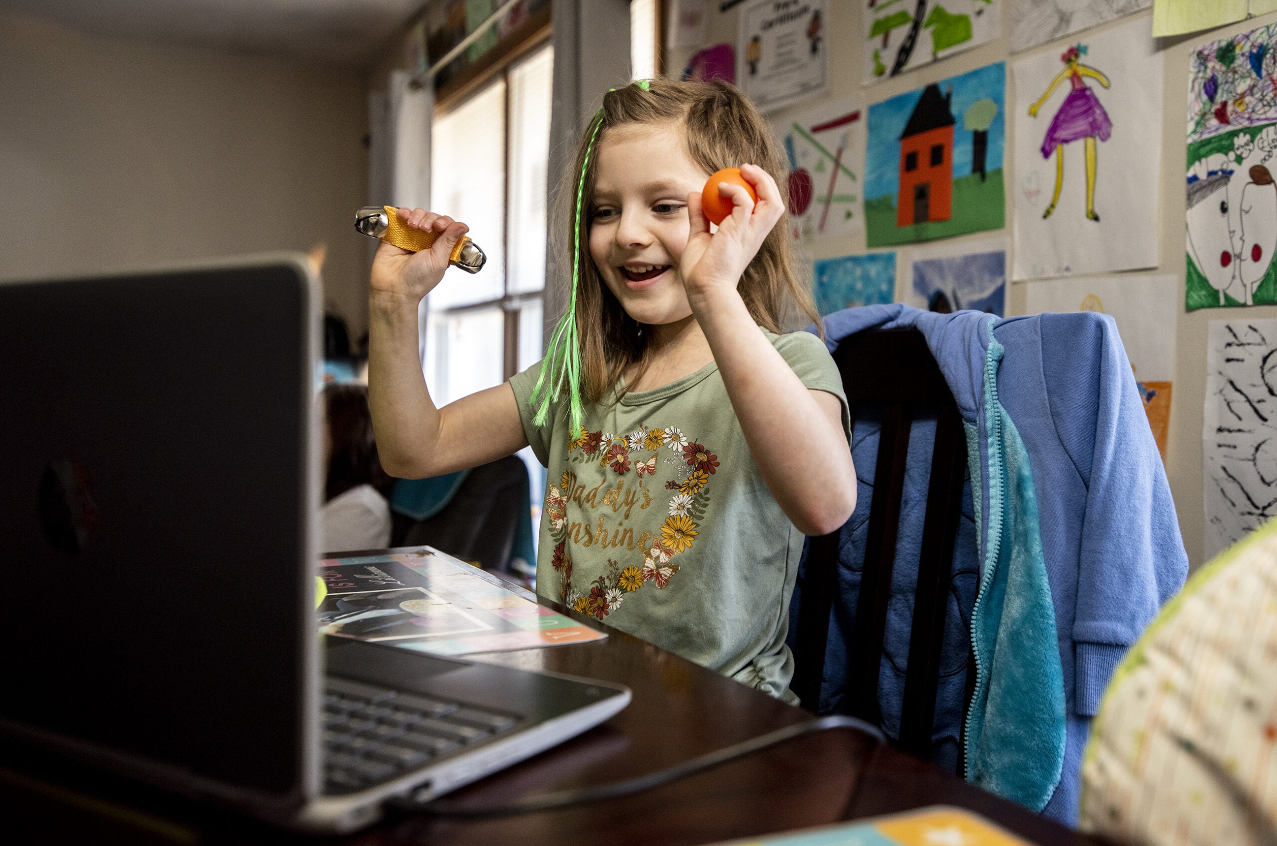 A student sits at a kitchen table in front of a laptop. In her raised hands, she holds bells and a percussion shaker.