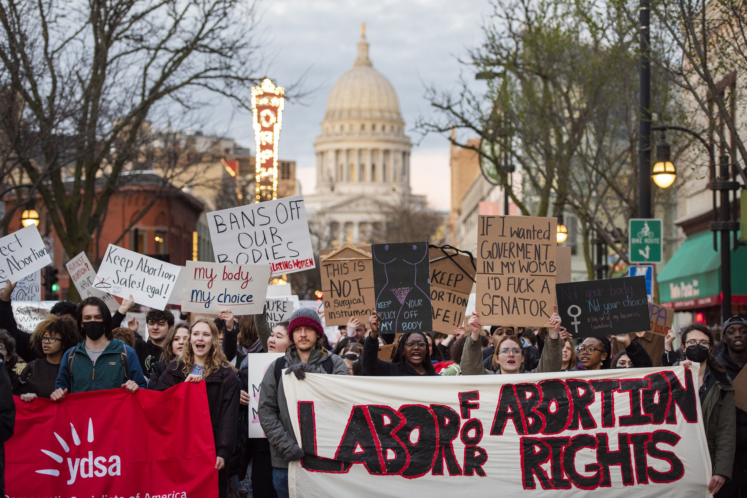 Protesters hold banners and raise signs as they march. The Capitol can be seen behind them.