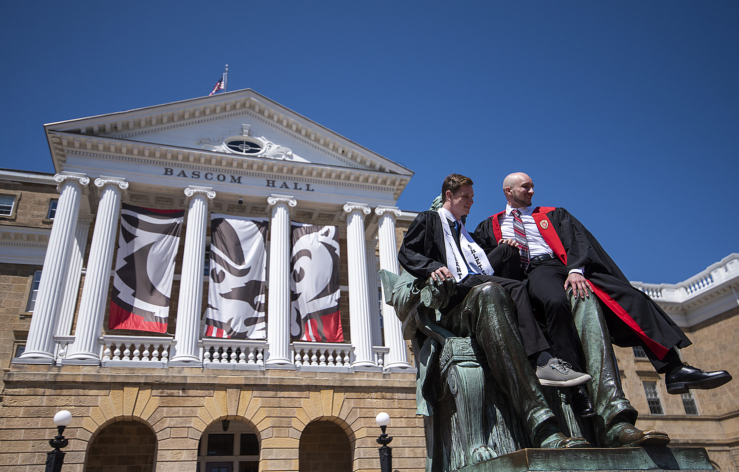 Two graduates in caps and gowns sit on the Abraham Lincoln statue at Bascom Hill at UW-Madison to take photos.