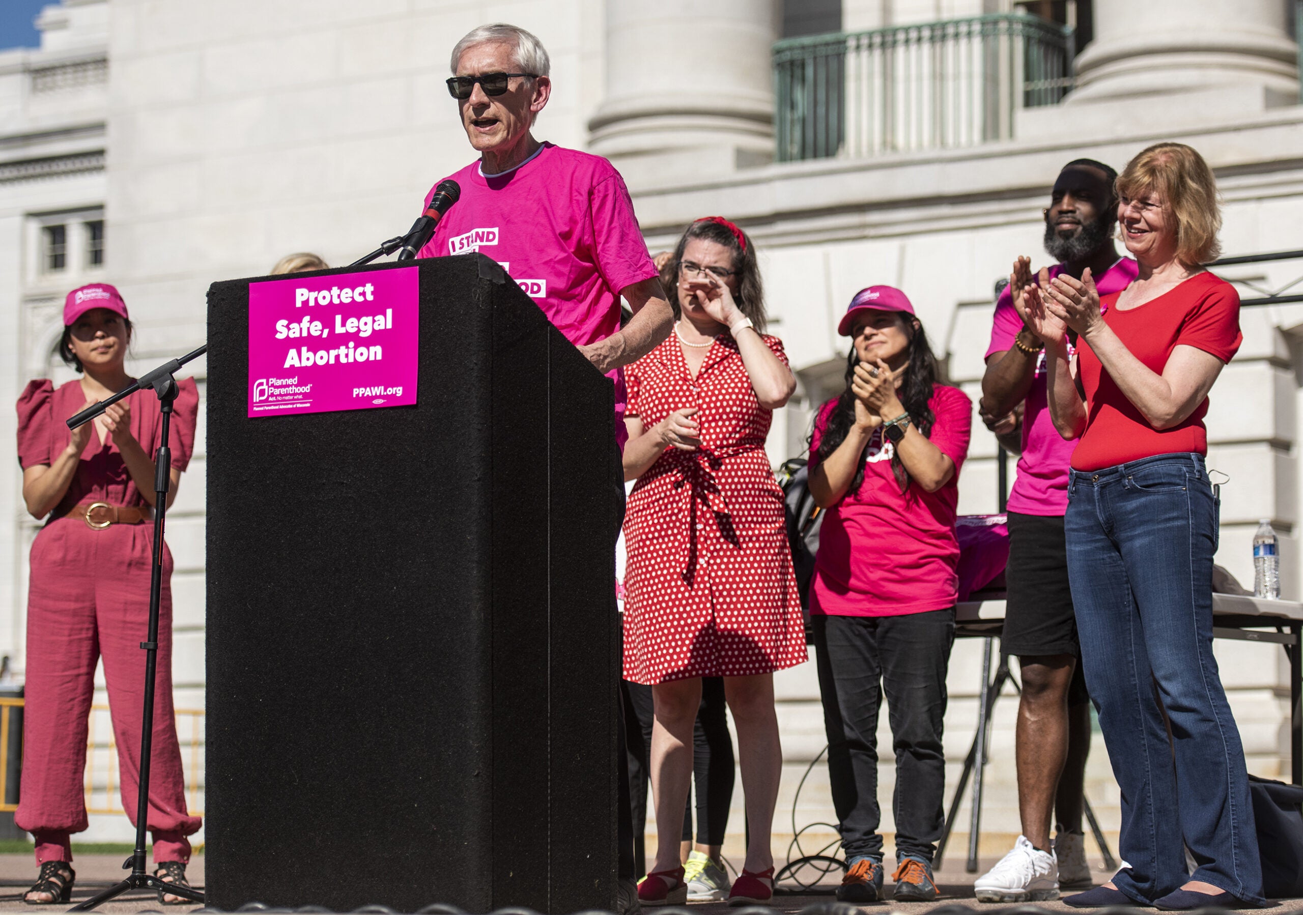 Gov. Evers speaks at a podium on the Wisconsin state Capitol steps. Sen. Tammy Baldwin applauds behind him as he speaks.