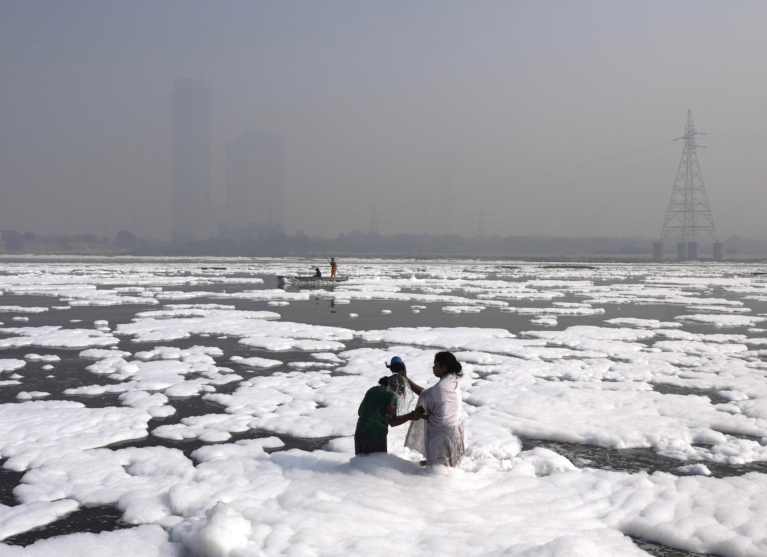 Two women in a polluted river next to smog-filled city