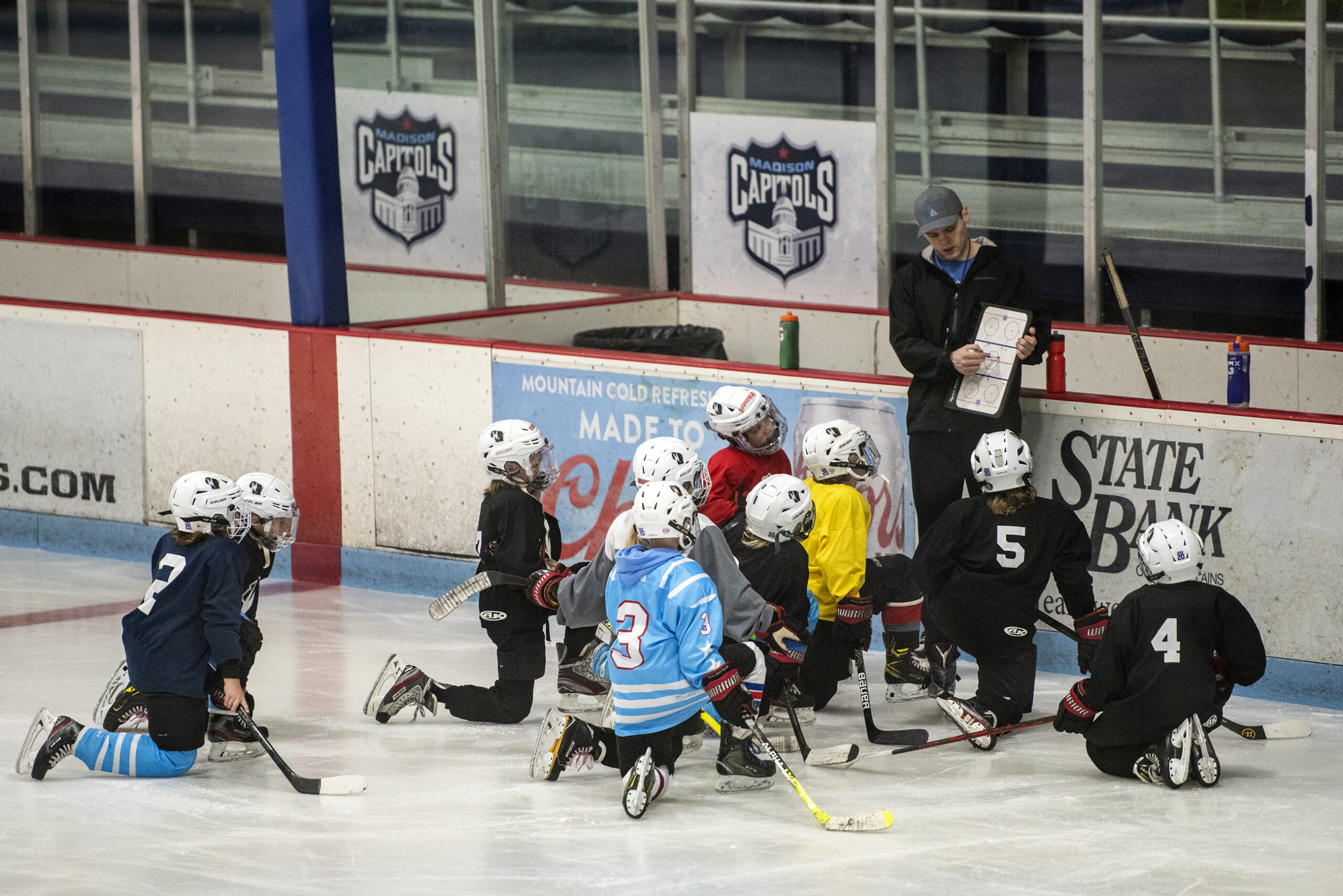 A group of youth hockey players kneel on the ice as a coach speaks to them.
