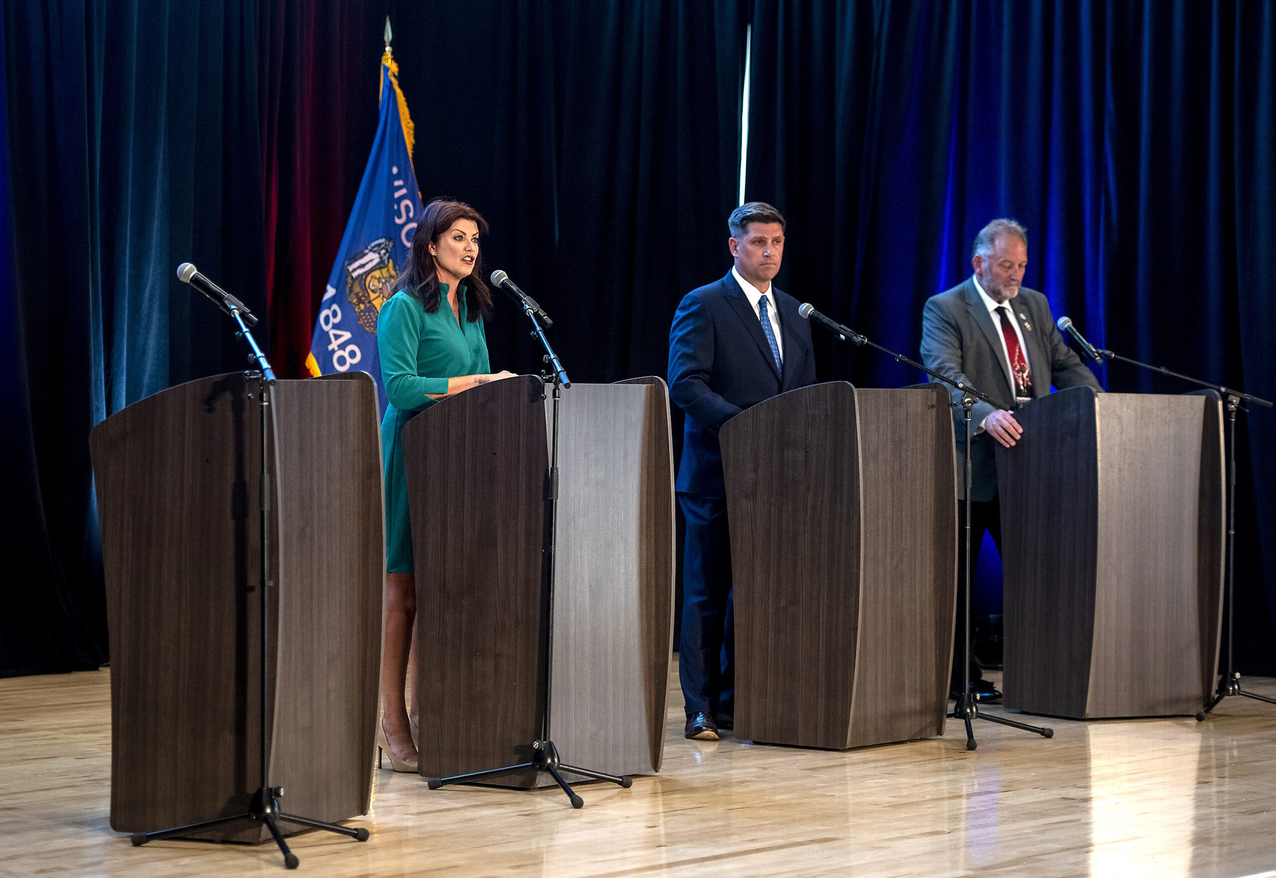 Three candidates stand at four podiums on stage.