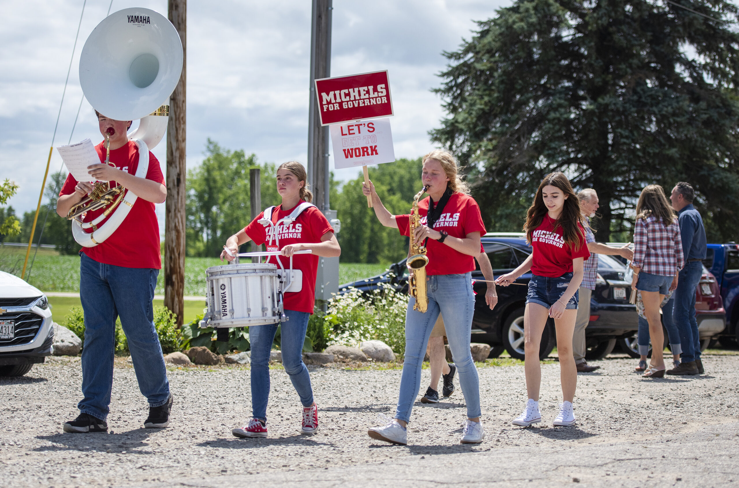 Musicians playing a sousaphone, snare drum and saxophone wear matching red shirts as they play outside of a golf club.