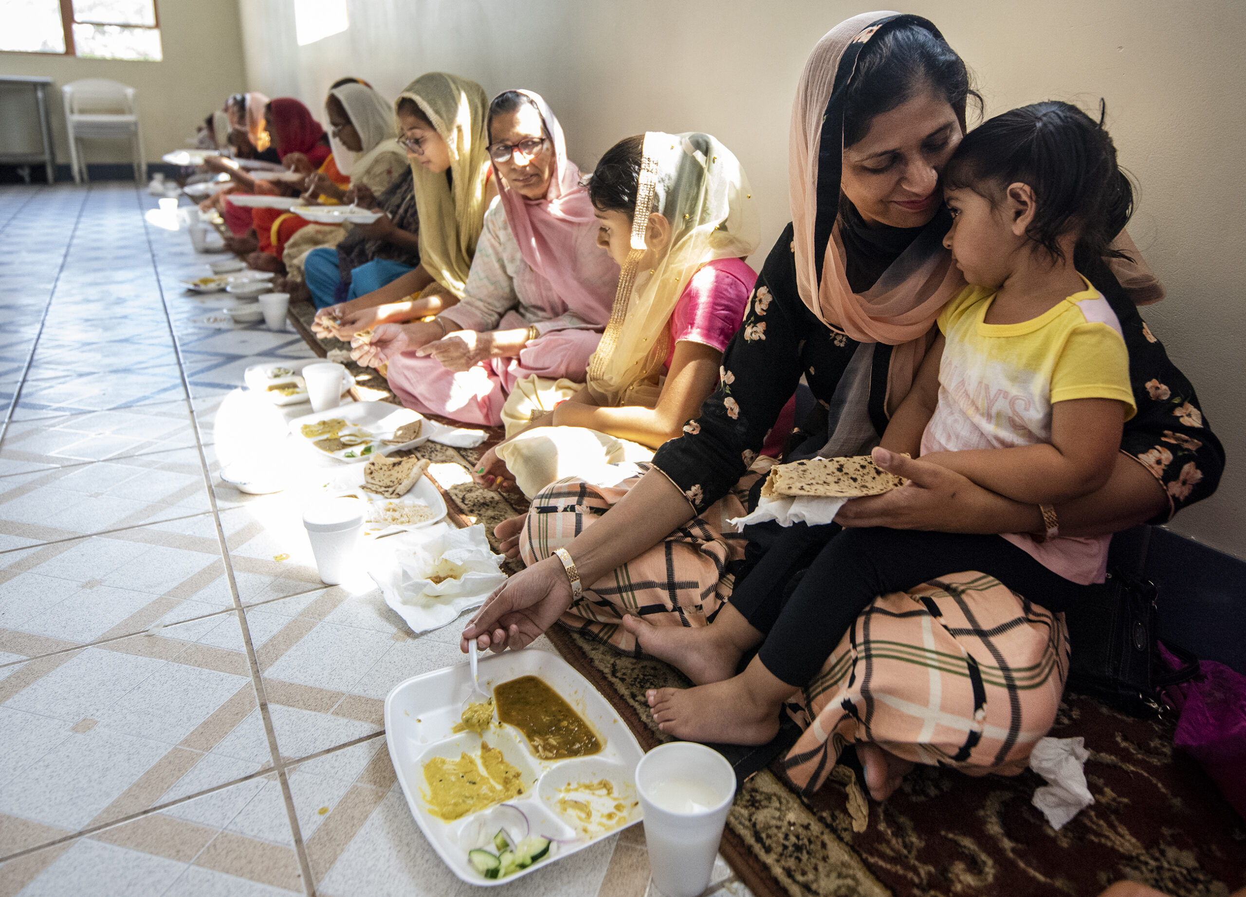 A woman holds a child as they sit on a rug to eat food.