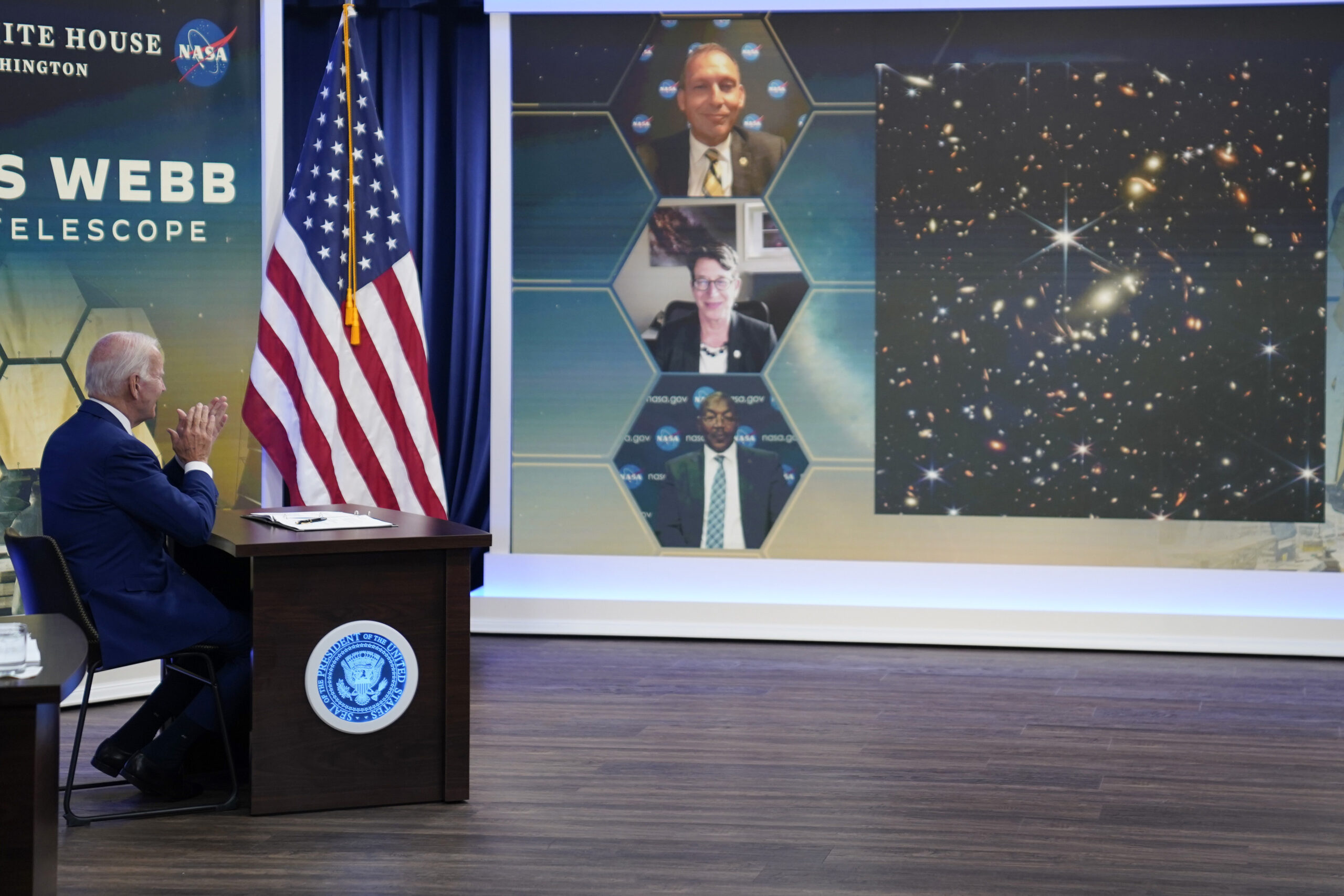 Joe Biden sits at a table and listens to NASA officials on a video screen