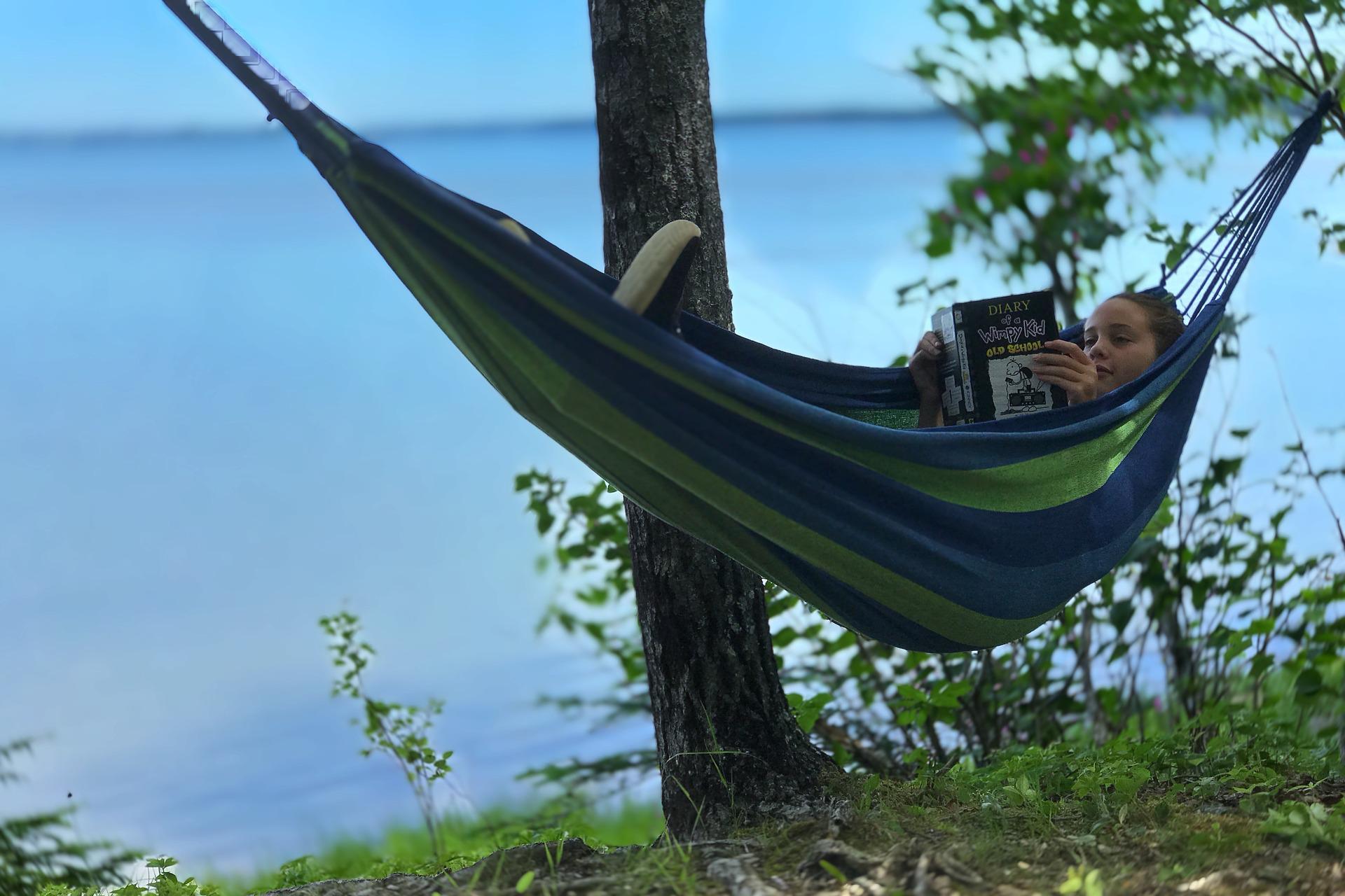 A kid reads a book in a hammock by a lake