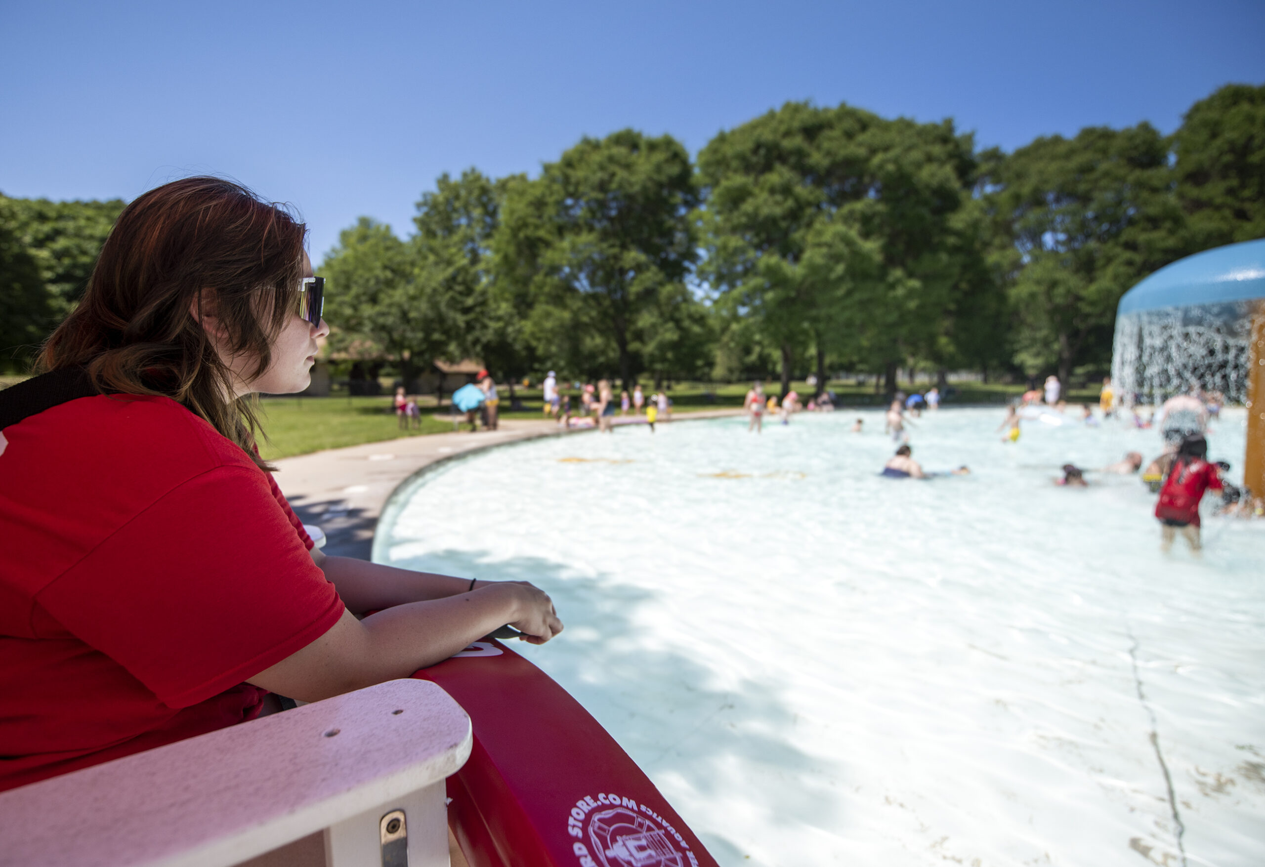 A lifeguard in red clothing watches a splash pool from a raised chair.