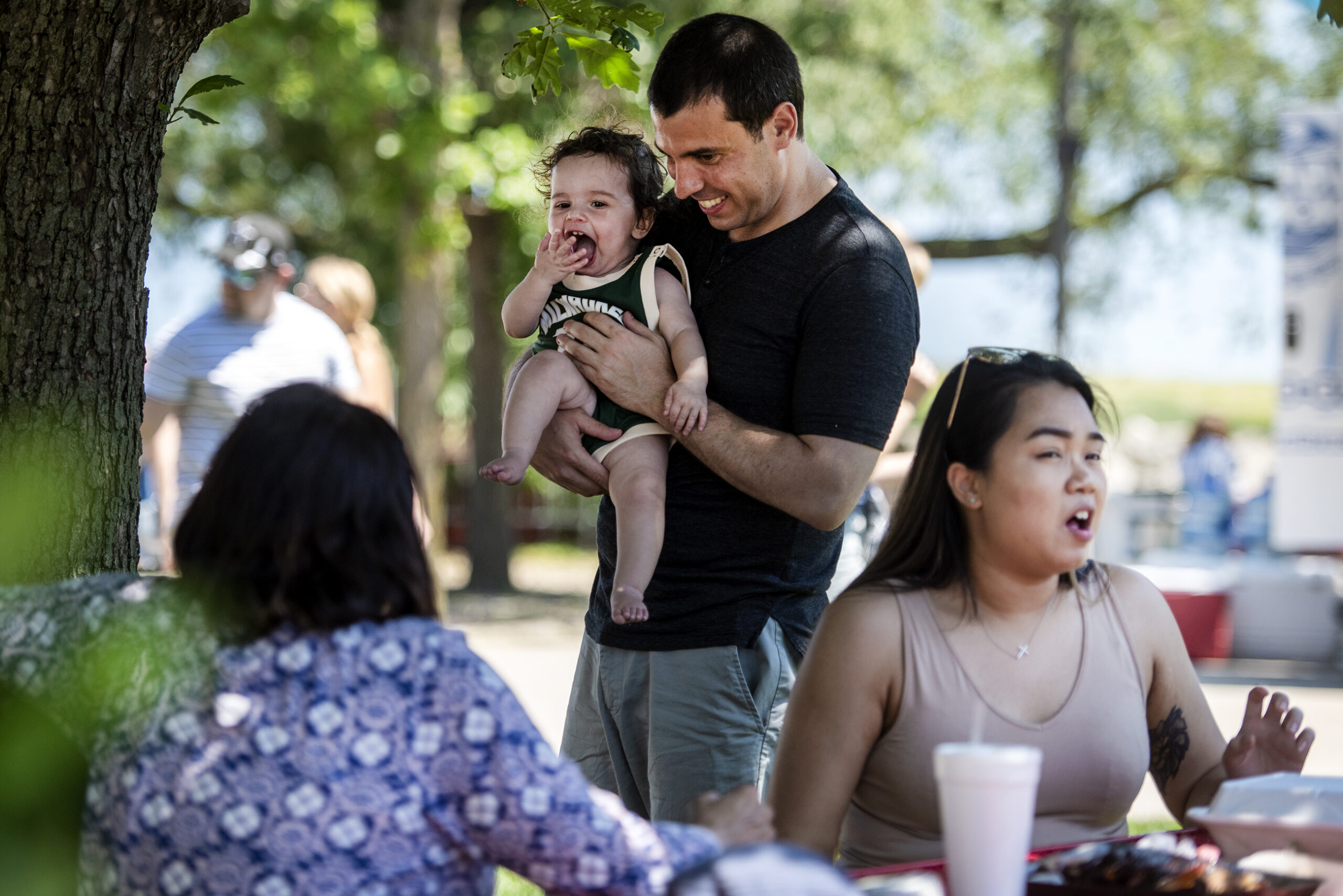 Alex Lasry holds his daughter as he speaks to people sitting at a picnic table.