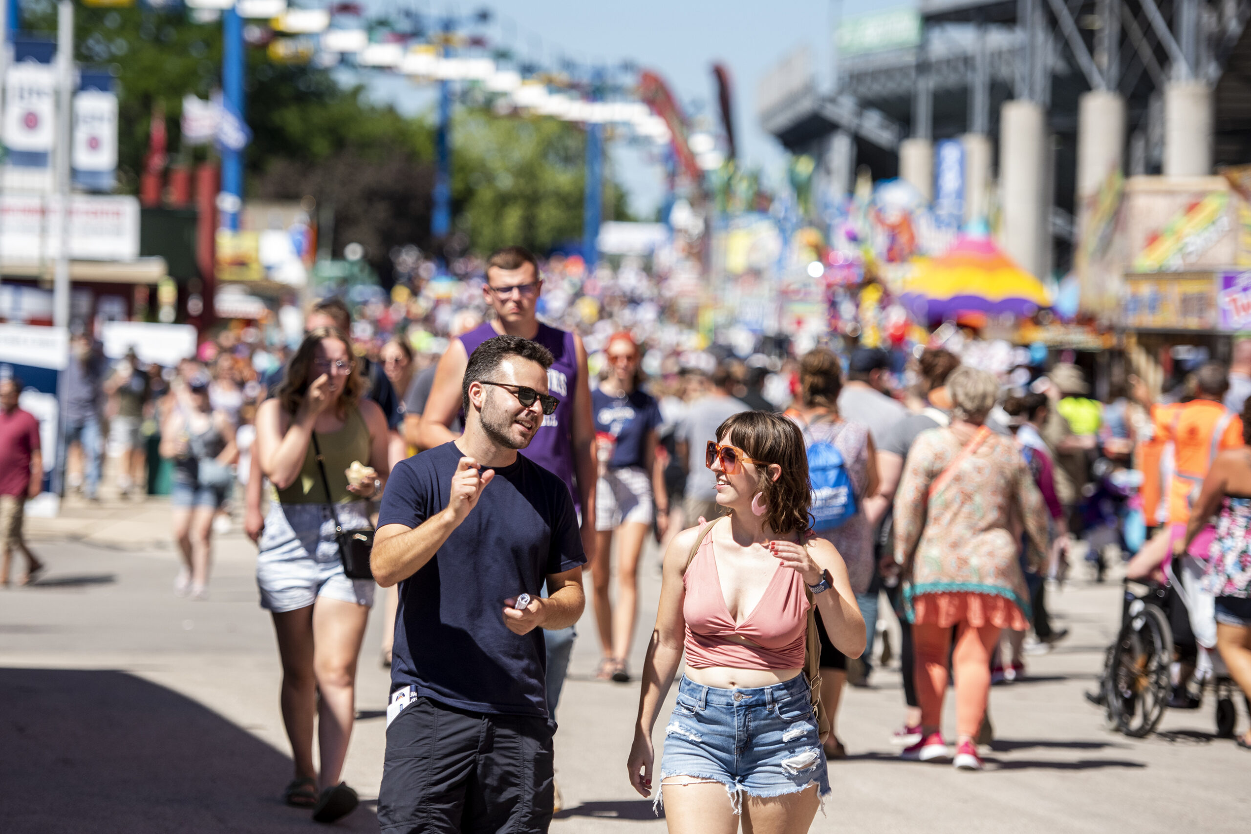 Two people walk together as a crowd walks behind them. Colorful fair booths are behind them.