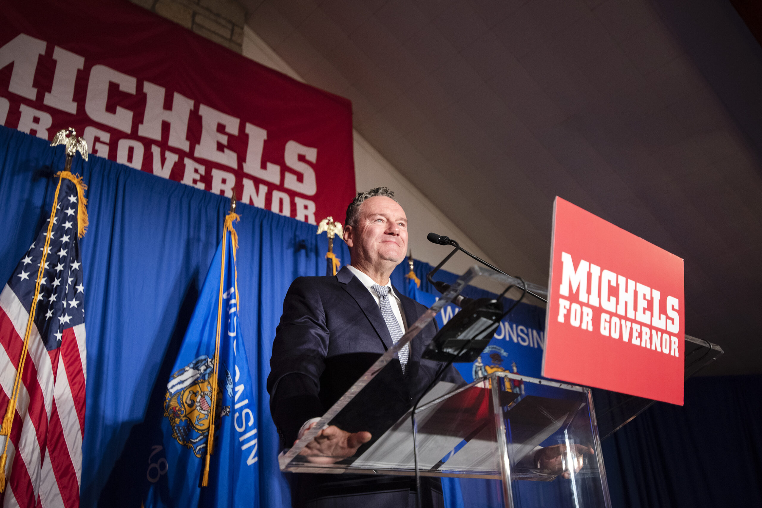 Tim Michels is surrounded by campaign signs as he addresses supporters on stage.