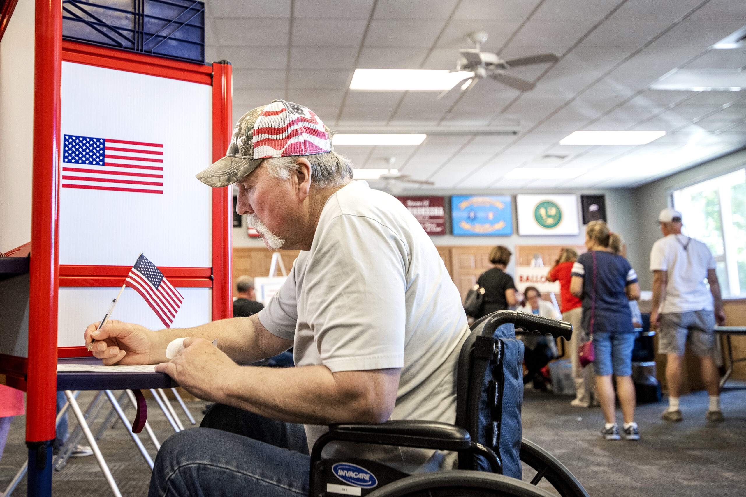 A man wears a hat with a U.S. flag pattern and writes with a pen with a flag attached as he votes at a booth with a flag decoration.
