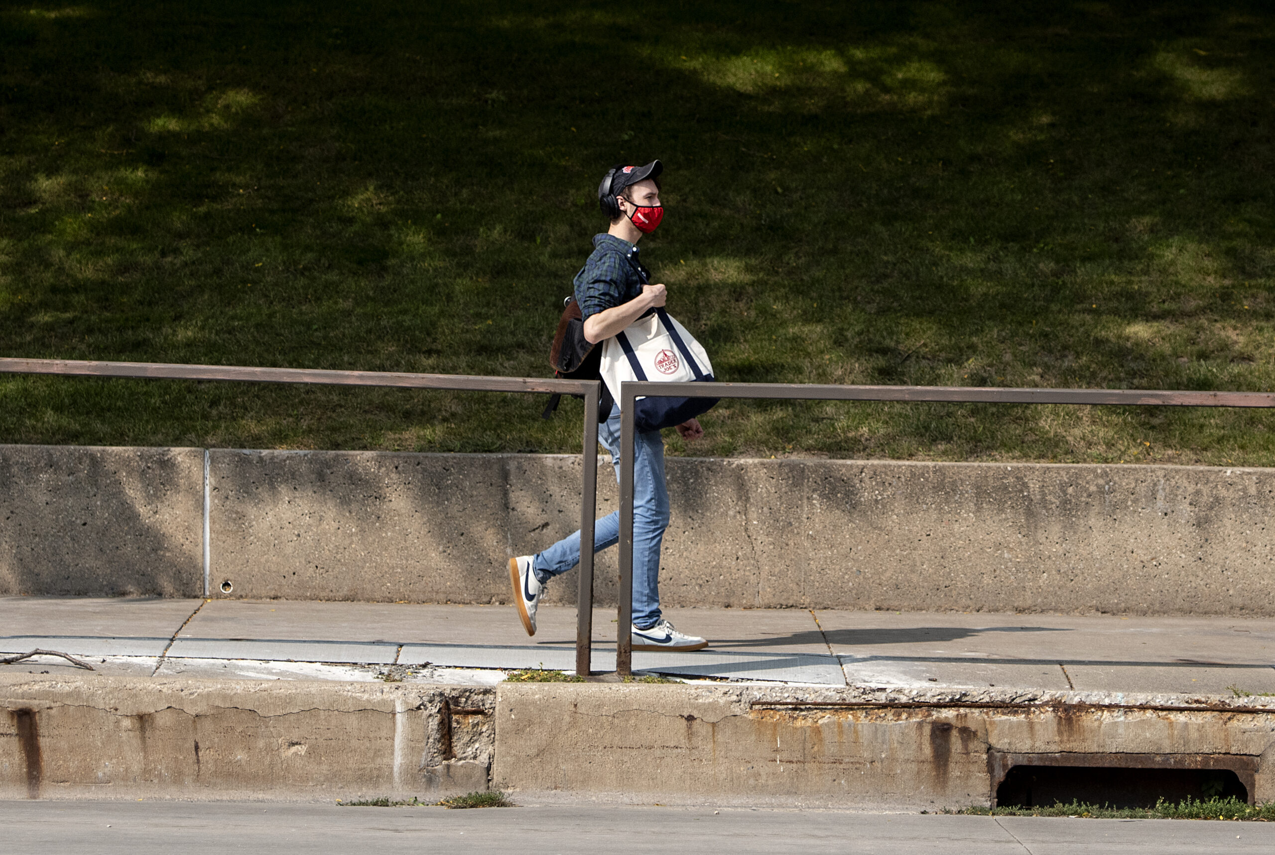 a pedestrian in a red face mask holds bags as he walks on the sidewalk on a sunny day