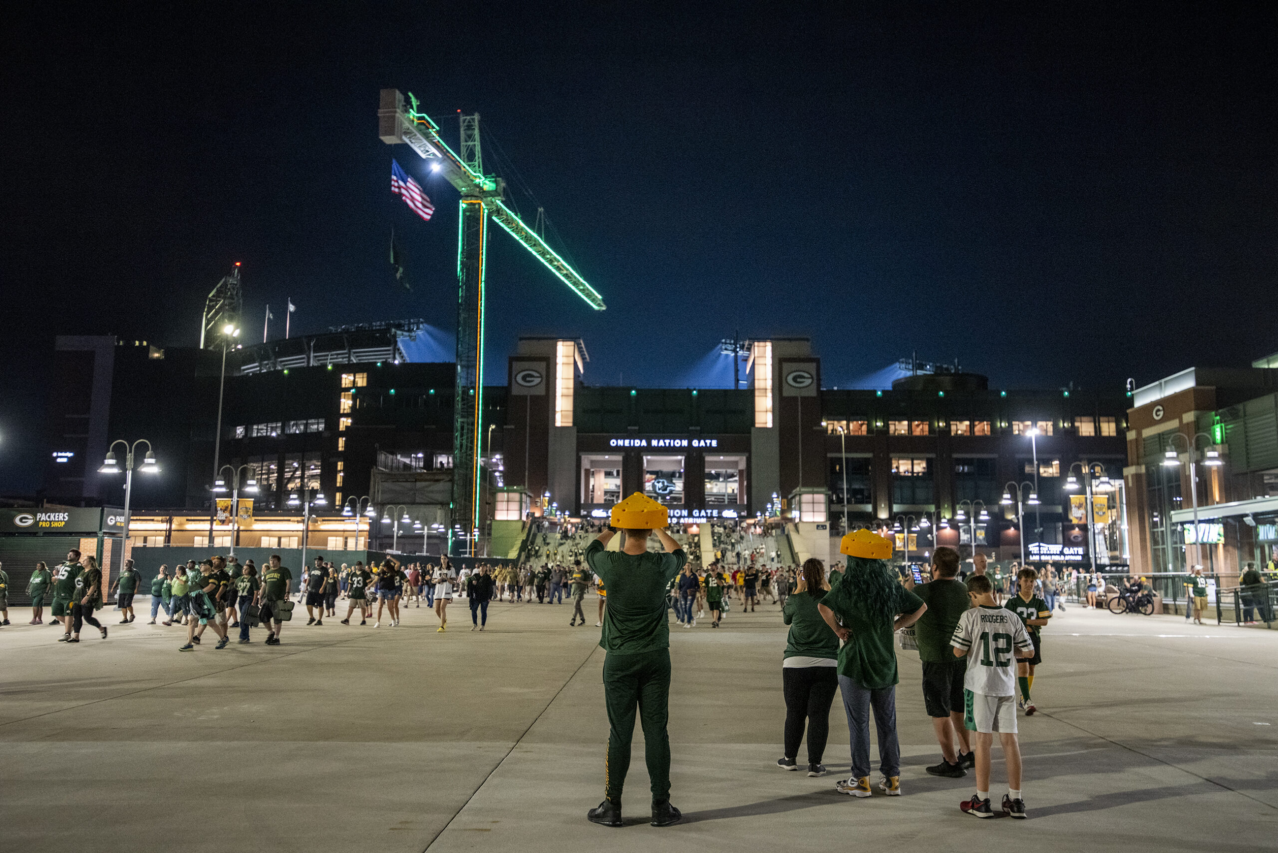 Fans wearing cheeseheads take photos of Lambeau Field against a night sky.