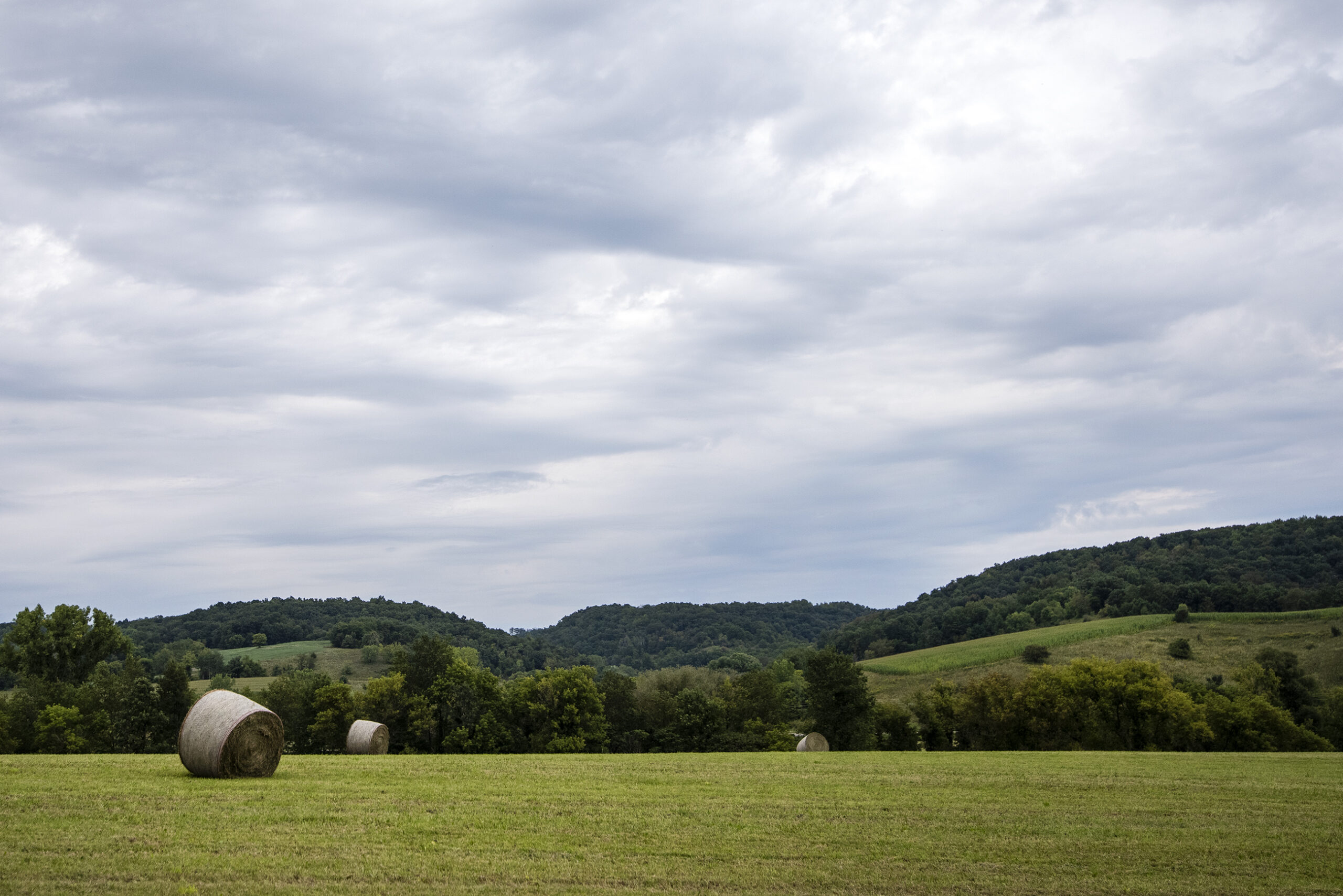 Bails of hay are seen in a flat field as hills rise in the distance.