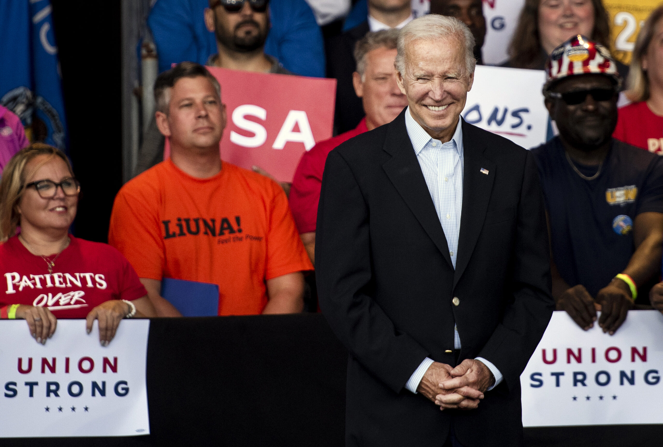 Pres. Biden smiles as he stands with his hands clasped. Supporters behind him hold signs that say, 