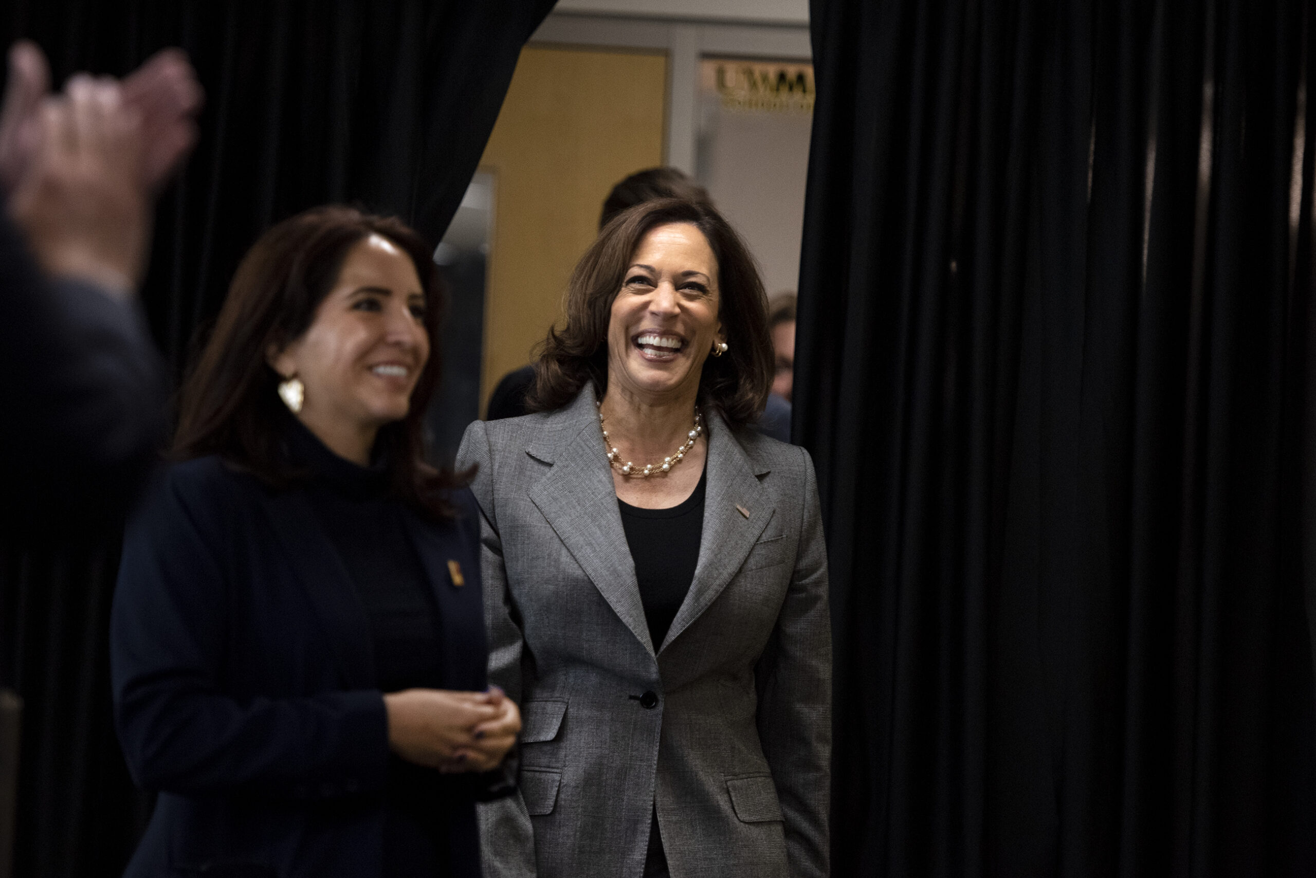 Vice President Kamala Harris enters out of a black curtain and smiles as she is greeted by people.