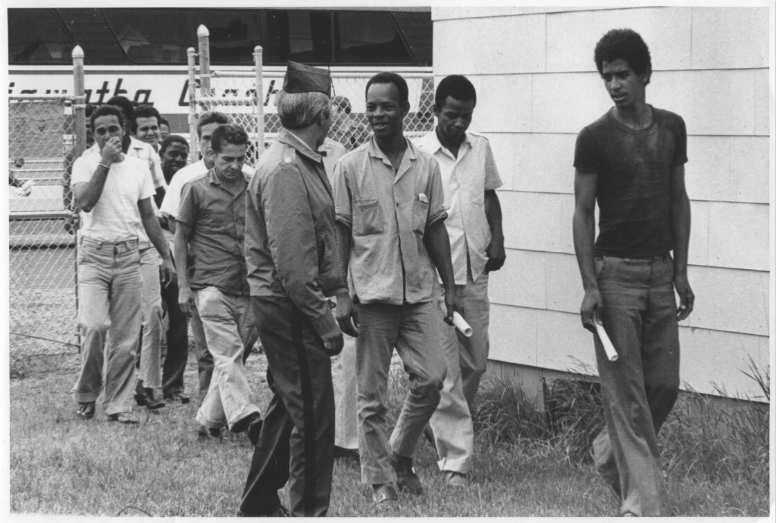 A black and white photograph featuring unidentified refugees walking together alongside a military personnel member.