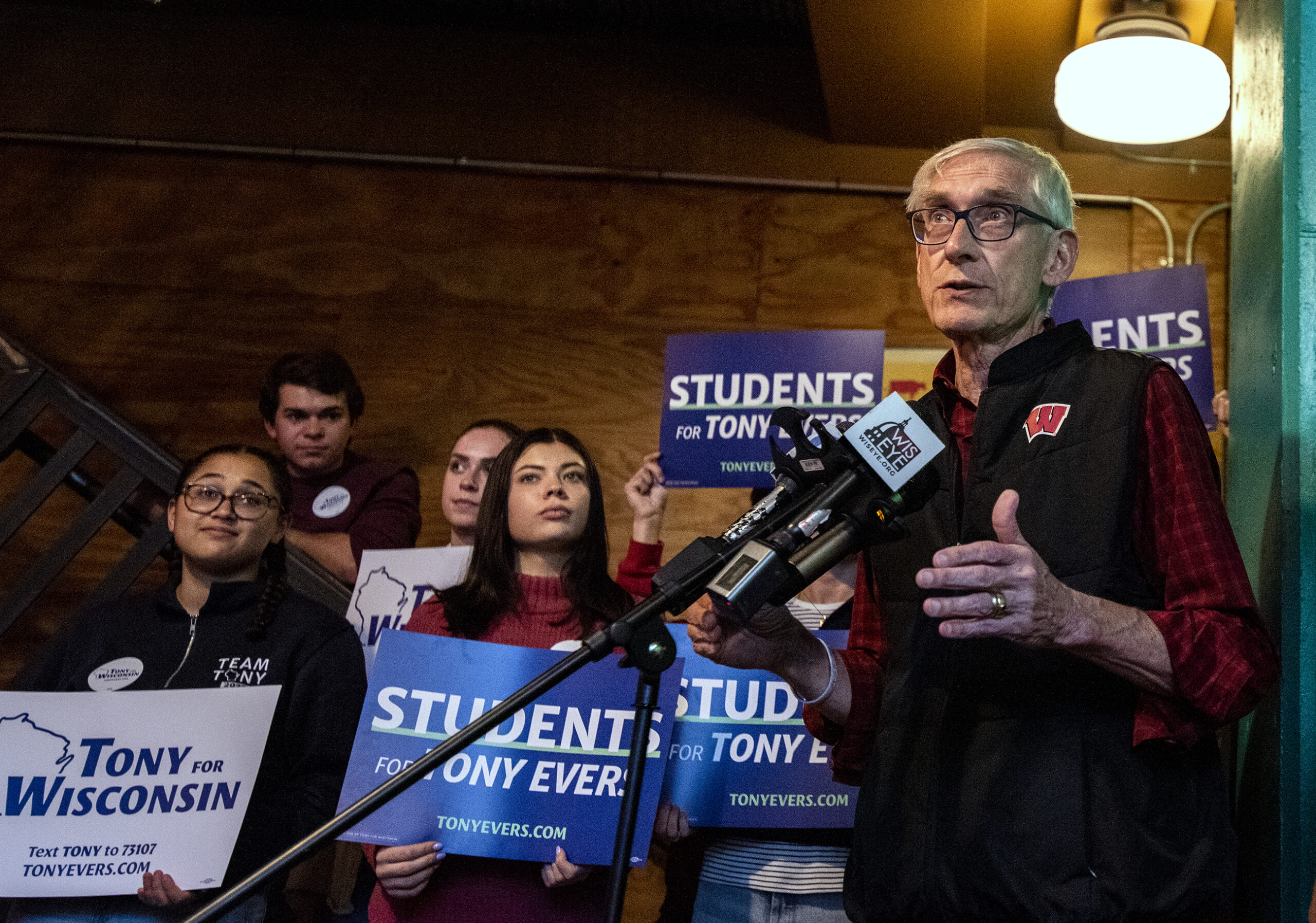 Gov. Tony Evers speaks into a microphone as students holding signs for his campaign stand behind him.