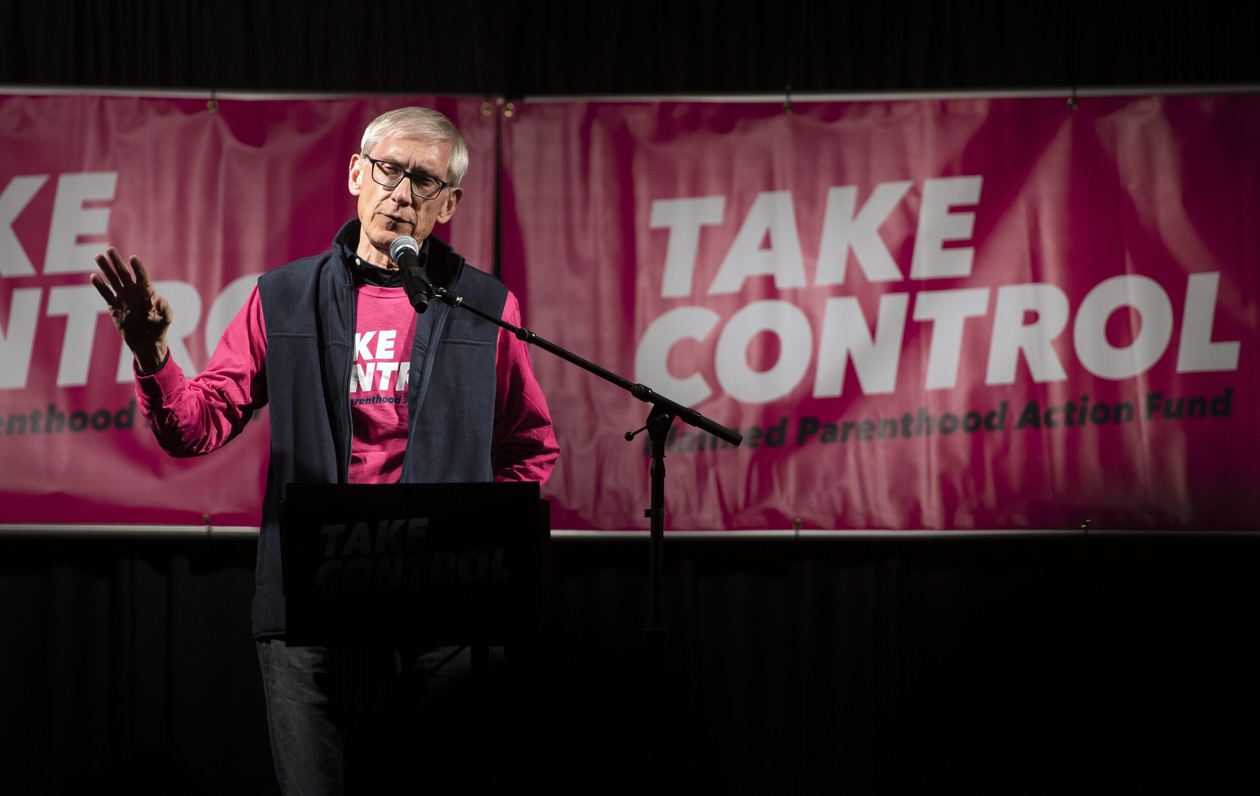Gov. Tony Evers speaks on stage in front of pink Planned Parenthood signs that say 