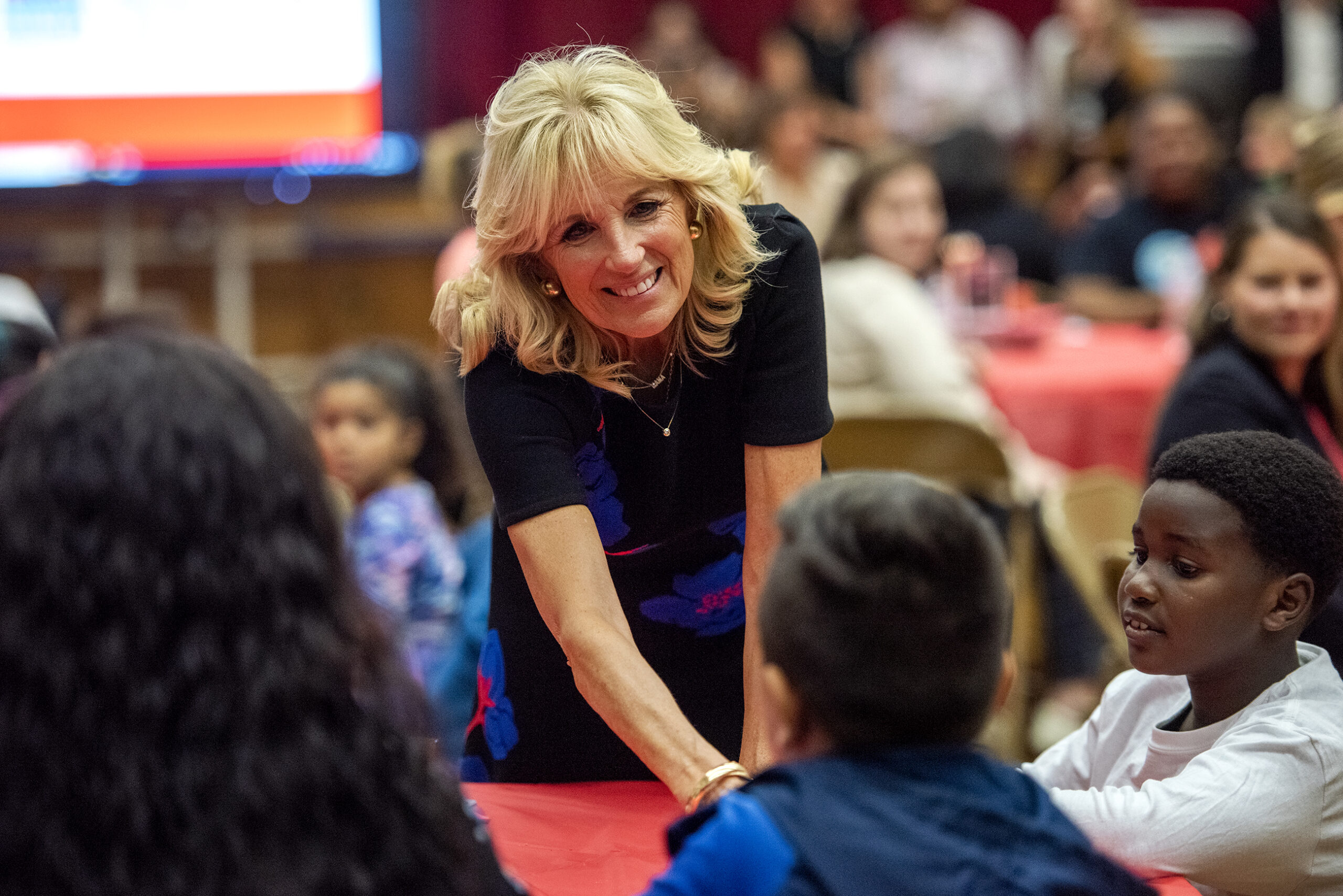 Jill Biden smiles as she leans over a table to shake hands with students and parents.