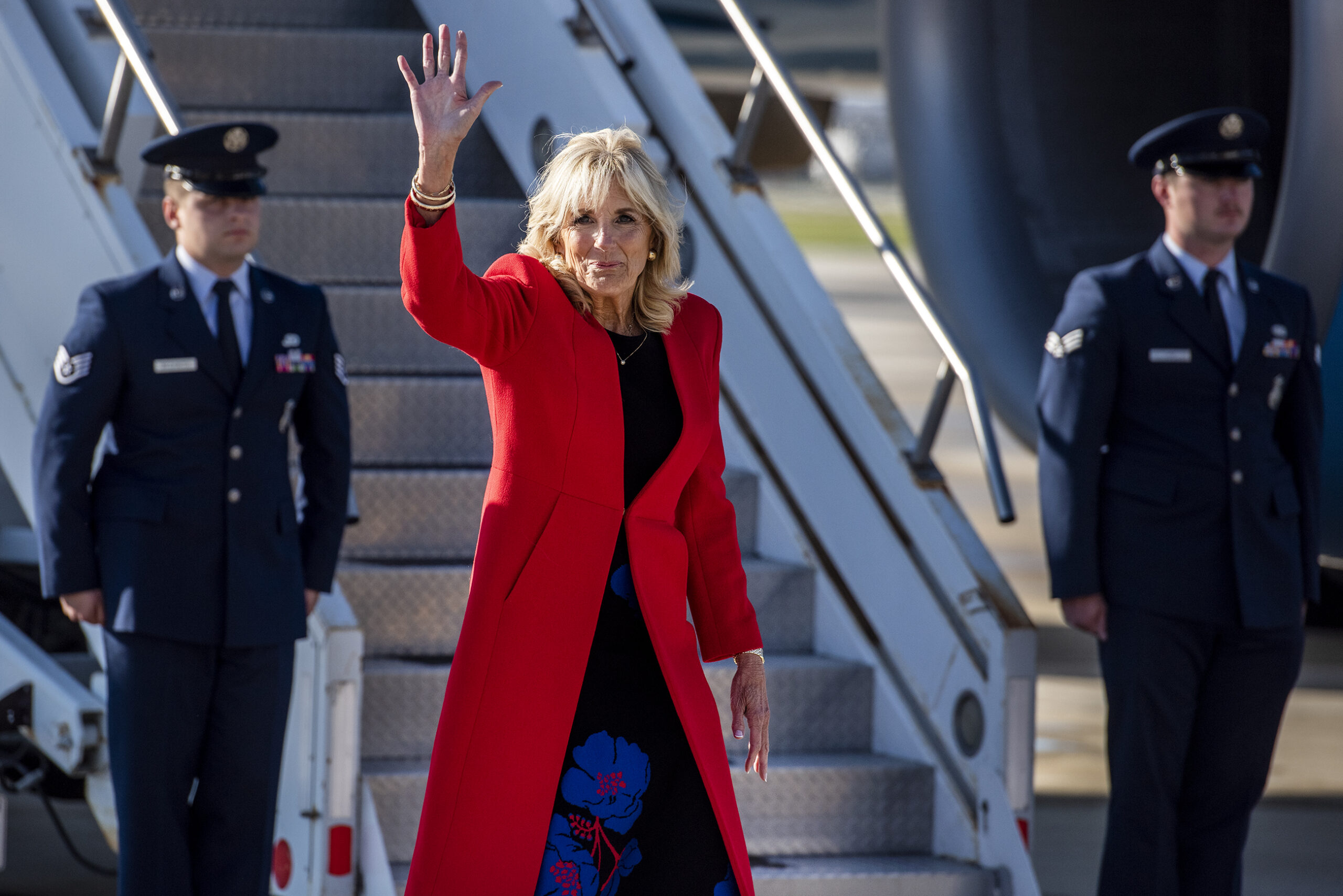 Jill Biden waves as she walks from the airplane to her vehicle. She wears a long red coat.
