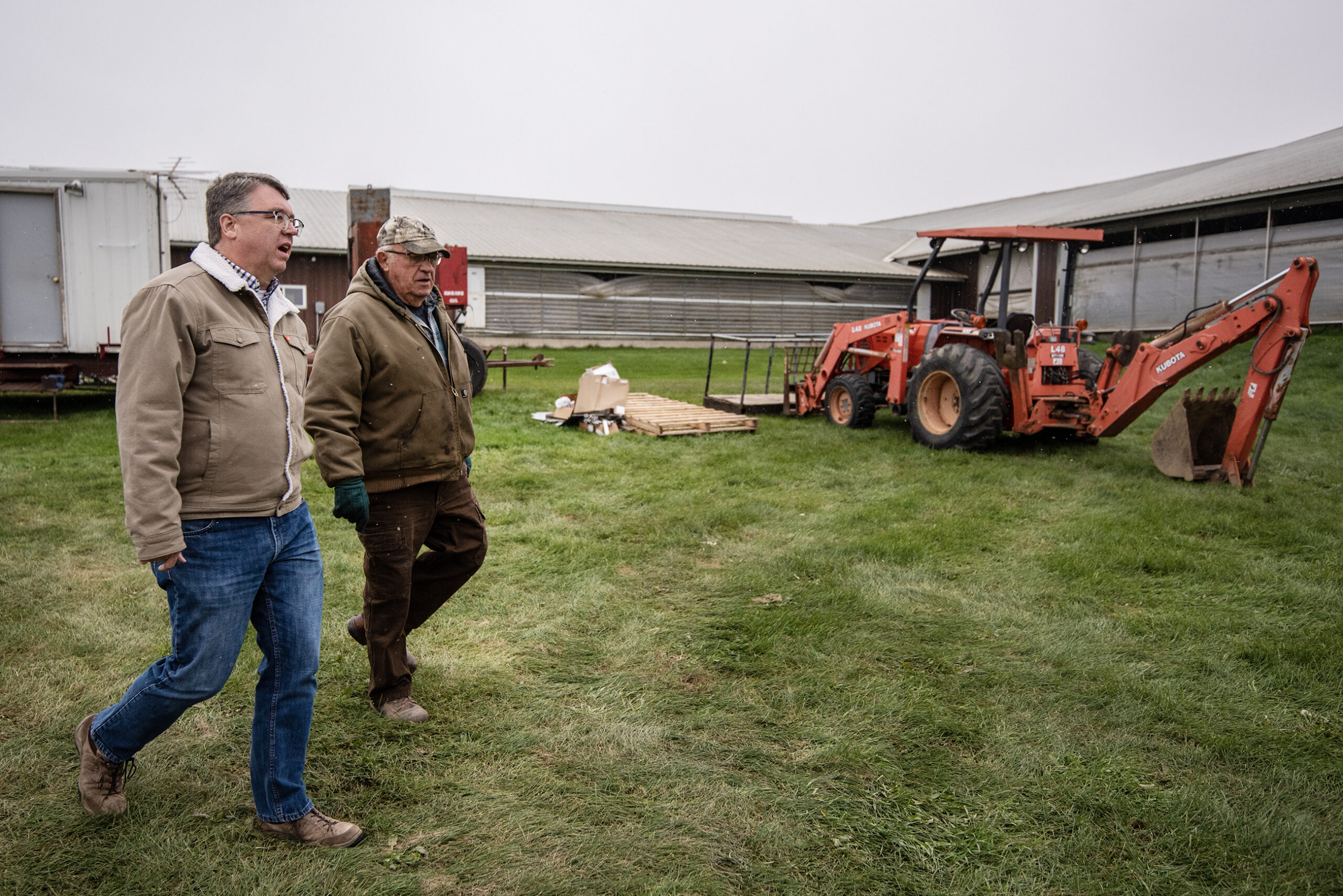 Two men walk together on green grass past farm equipment.