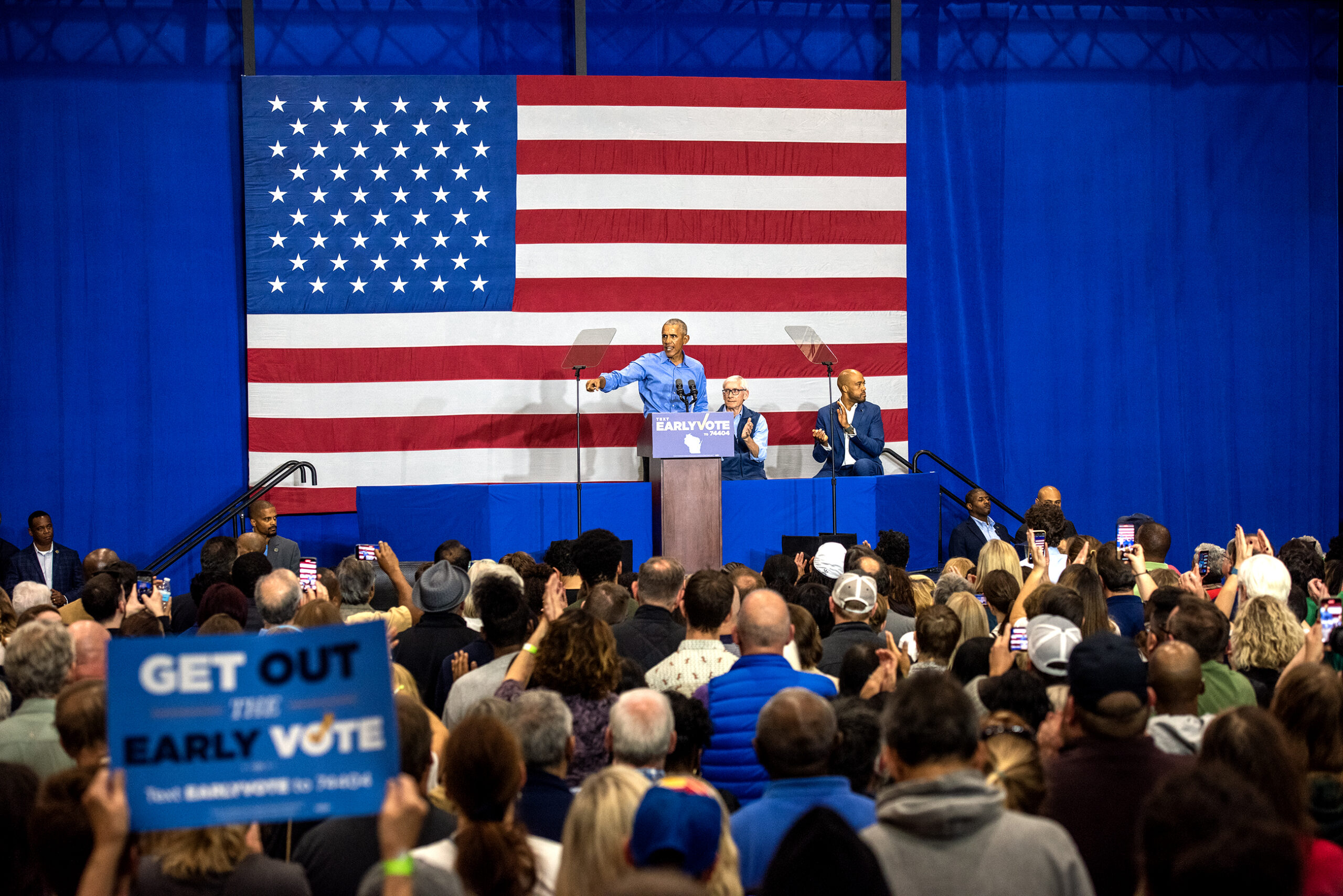 Former President Barack Obama stands in front of a large U.S. flag while he speaks in front of a sea of supporters.