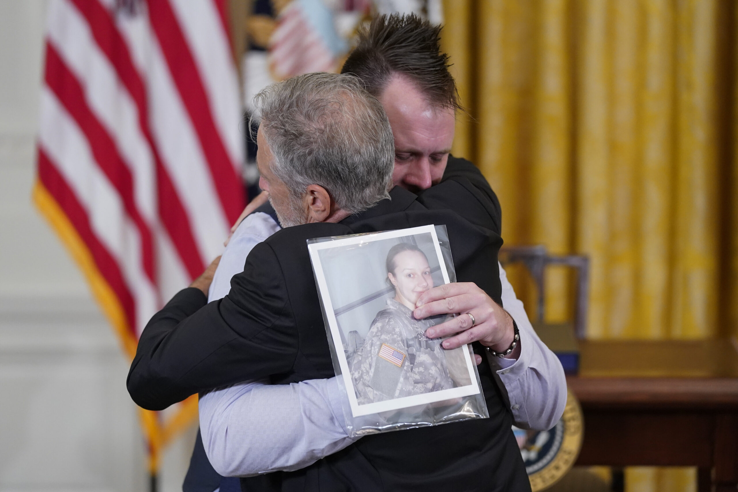 Comedian Jon Stewart hugs the husband of a veteran, whose picture the husband is holding