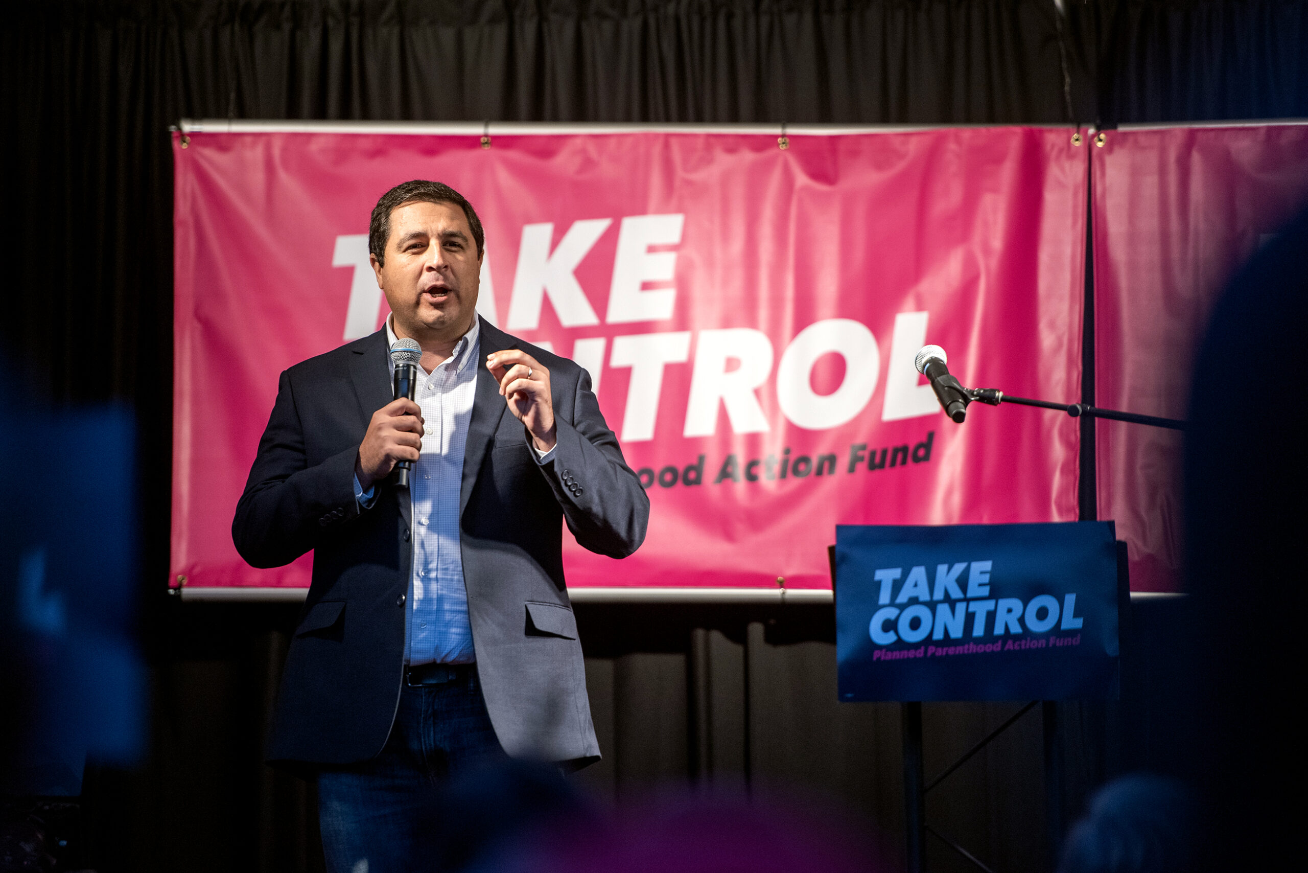 Attorney General Josh Kaul stands in front of a pink Planned Parenthood banner that says "take control" as he speaks into a microphone on stage.