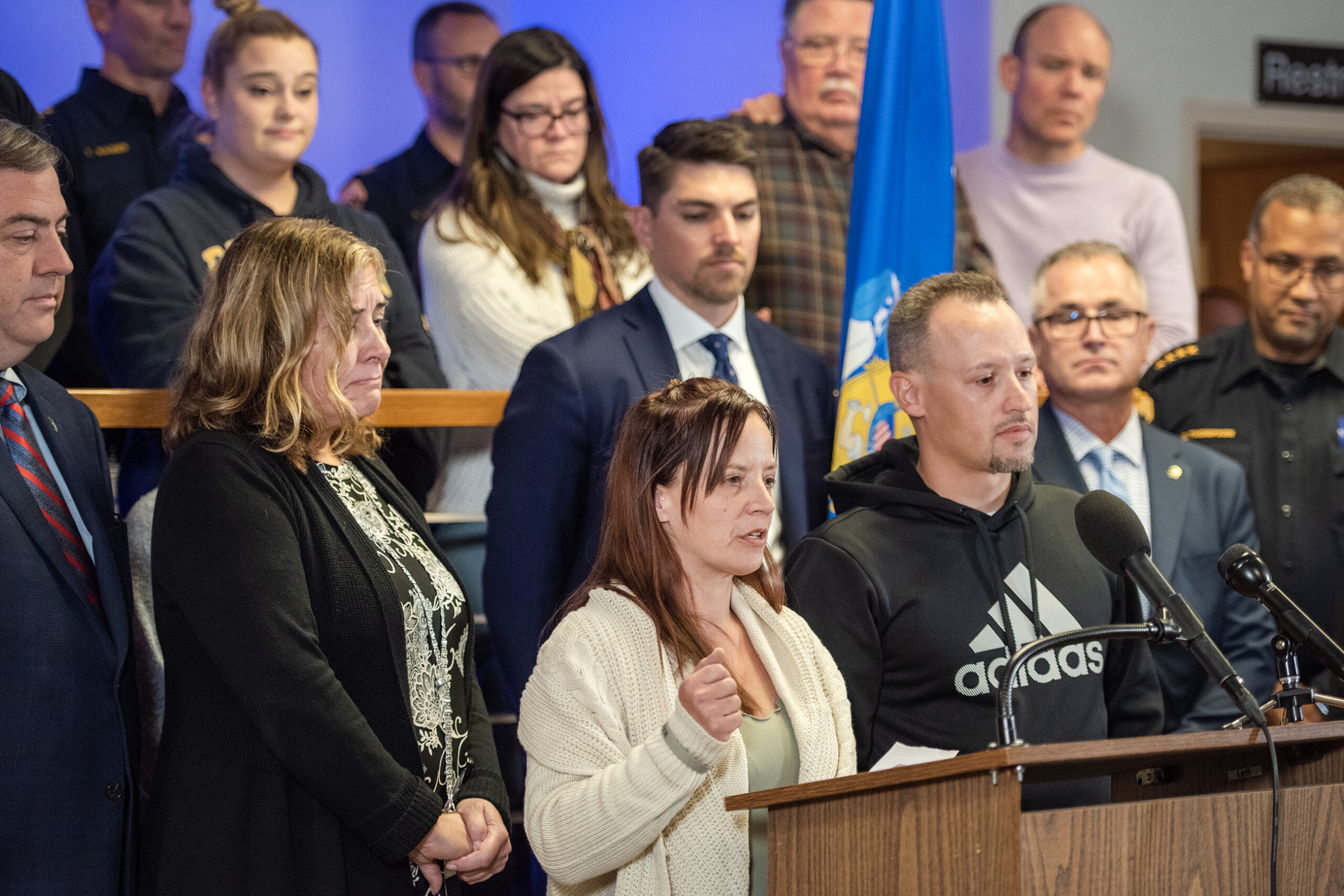 A woman is surrounded by other victims families and law enforcement as she speaks at a podium.