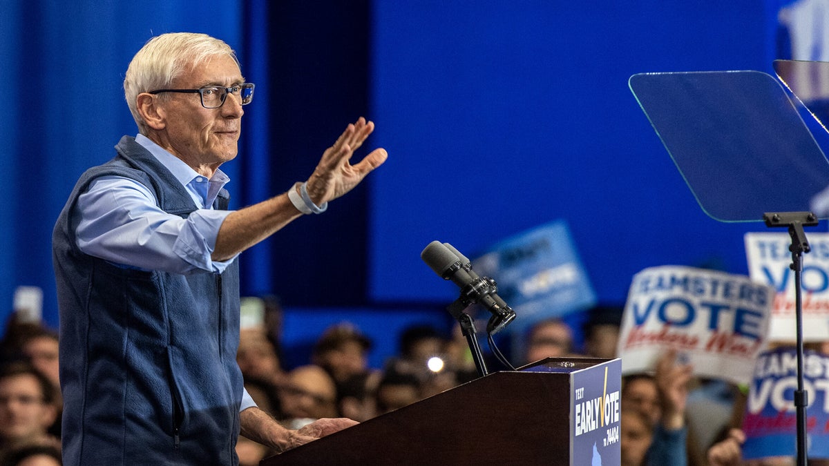 Gov. Tony Evers stands behind a podium and raises his hand to wave at a crowd.