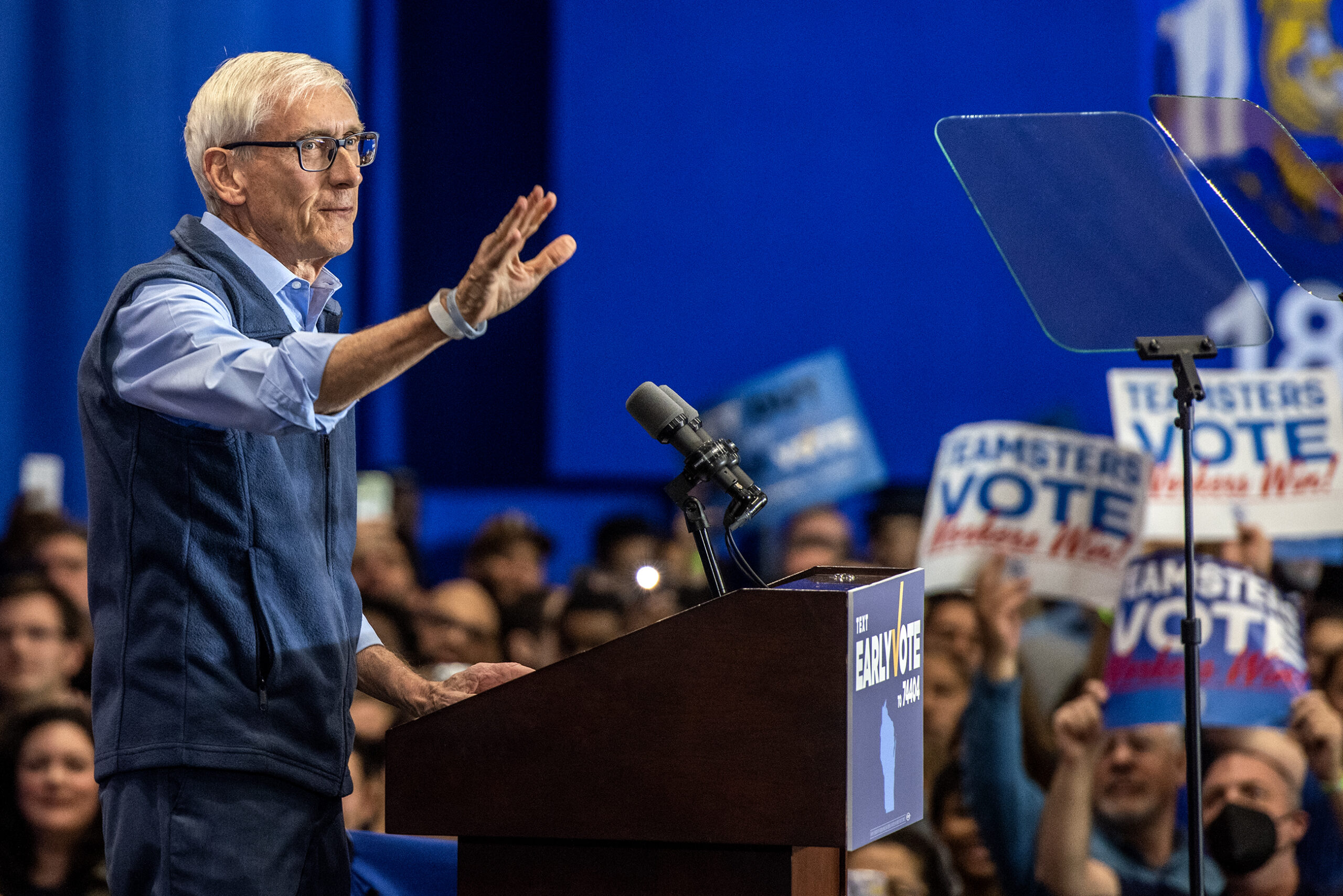 Gov. Tony Evers stands behind a podium and raises his hand to wave at a crowd.