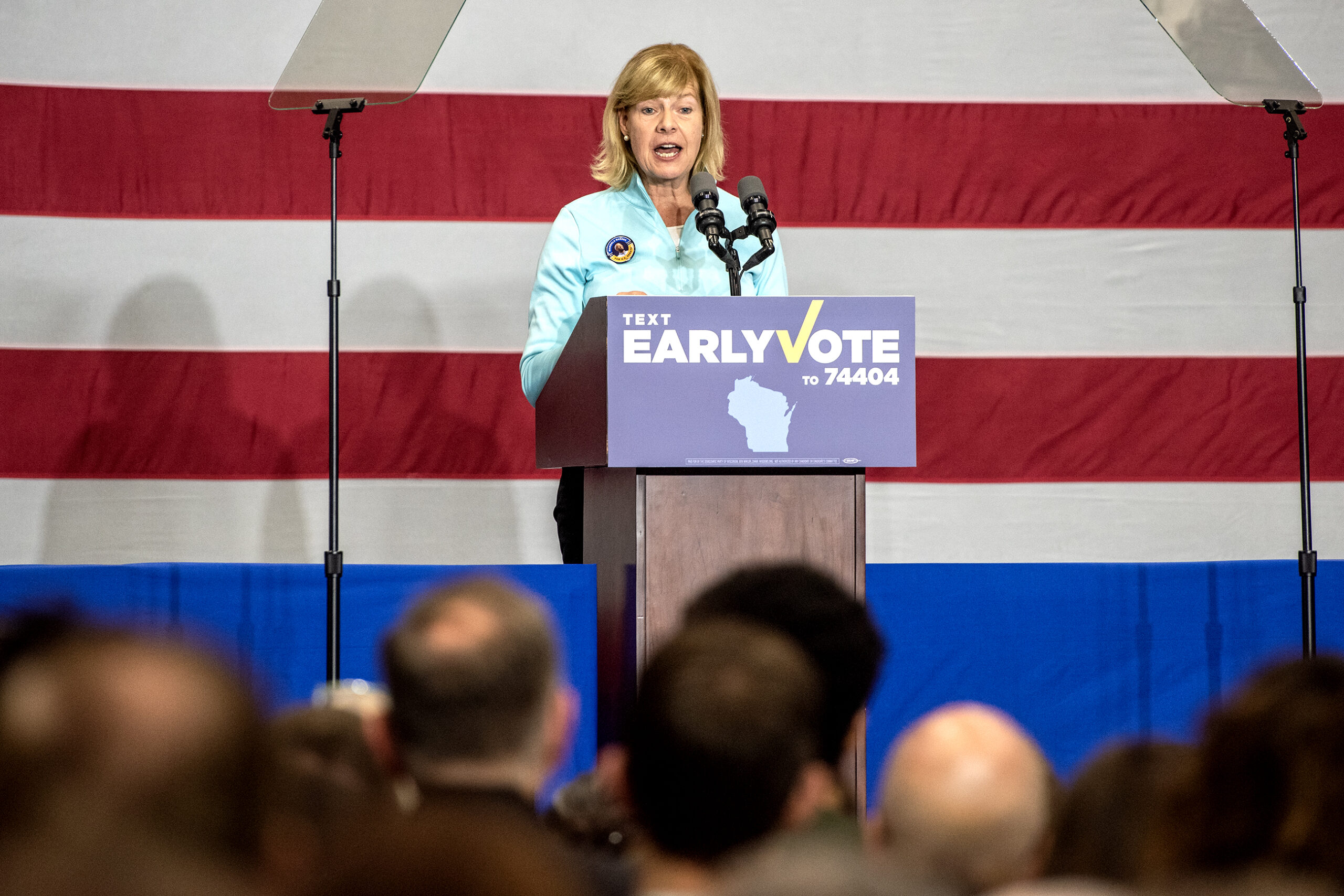 Sen. Tammy Baldwin speaks in front of a U.S. flag background. Attendees can be seen in the foreground.