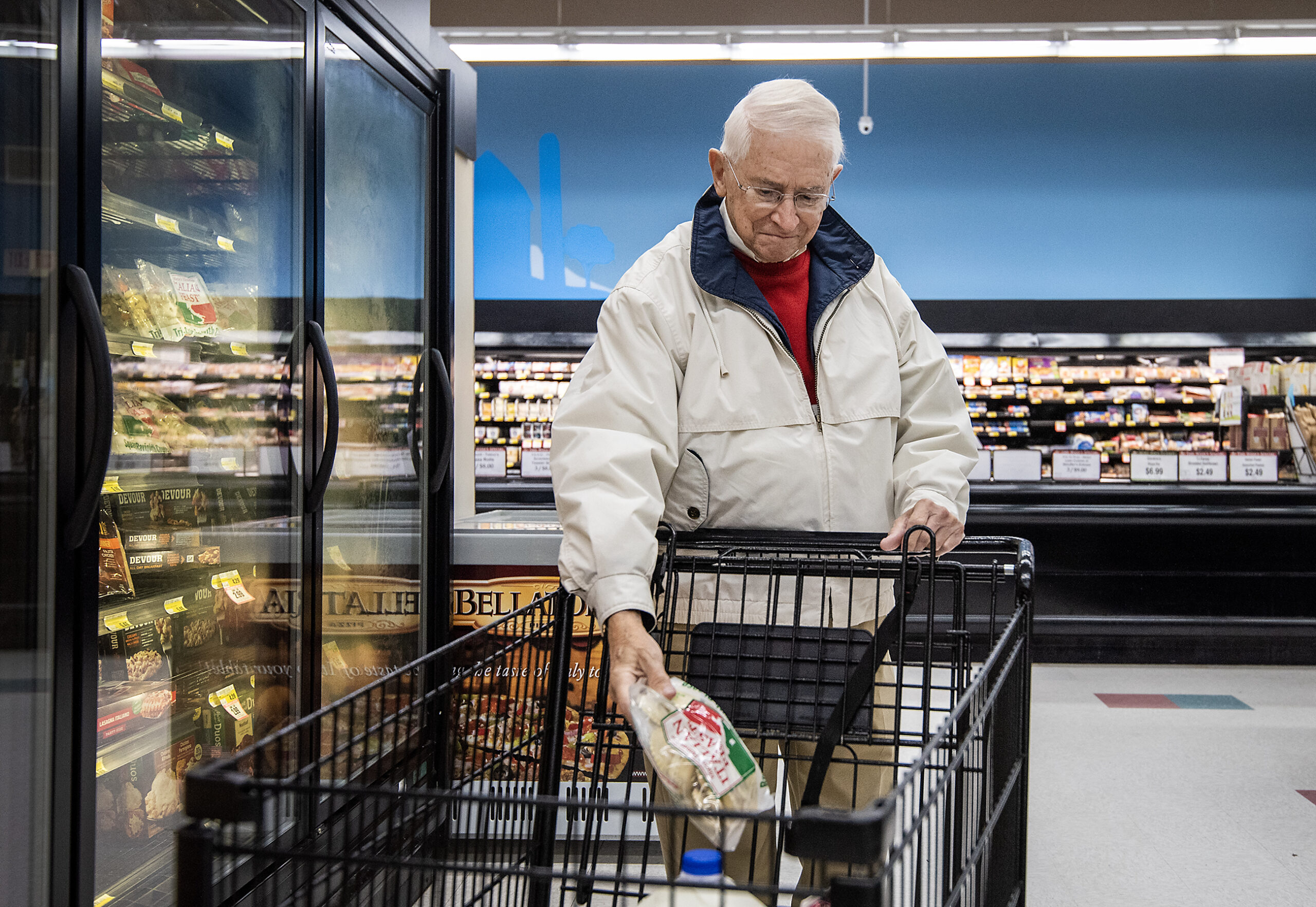 A man in a jacket puts a package into a cart in the frozen food aisle.