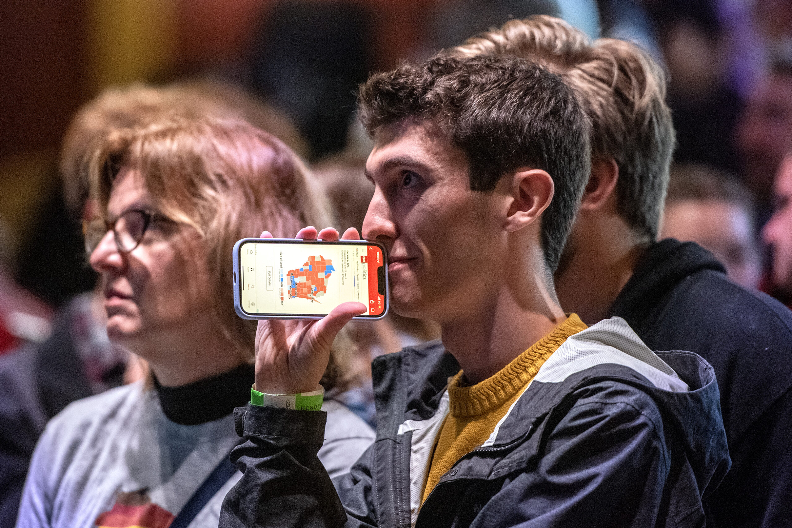 Attendees look up at a screen with pensive expressions. A phone screen shows a map of Wisconsin covered in red and blue sections, indicating election results.