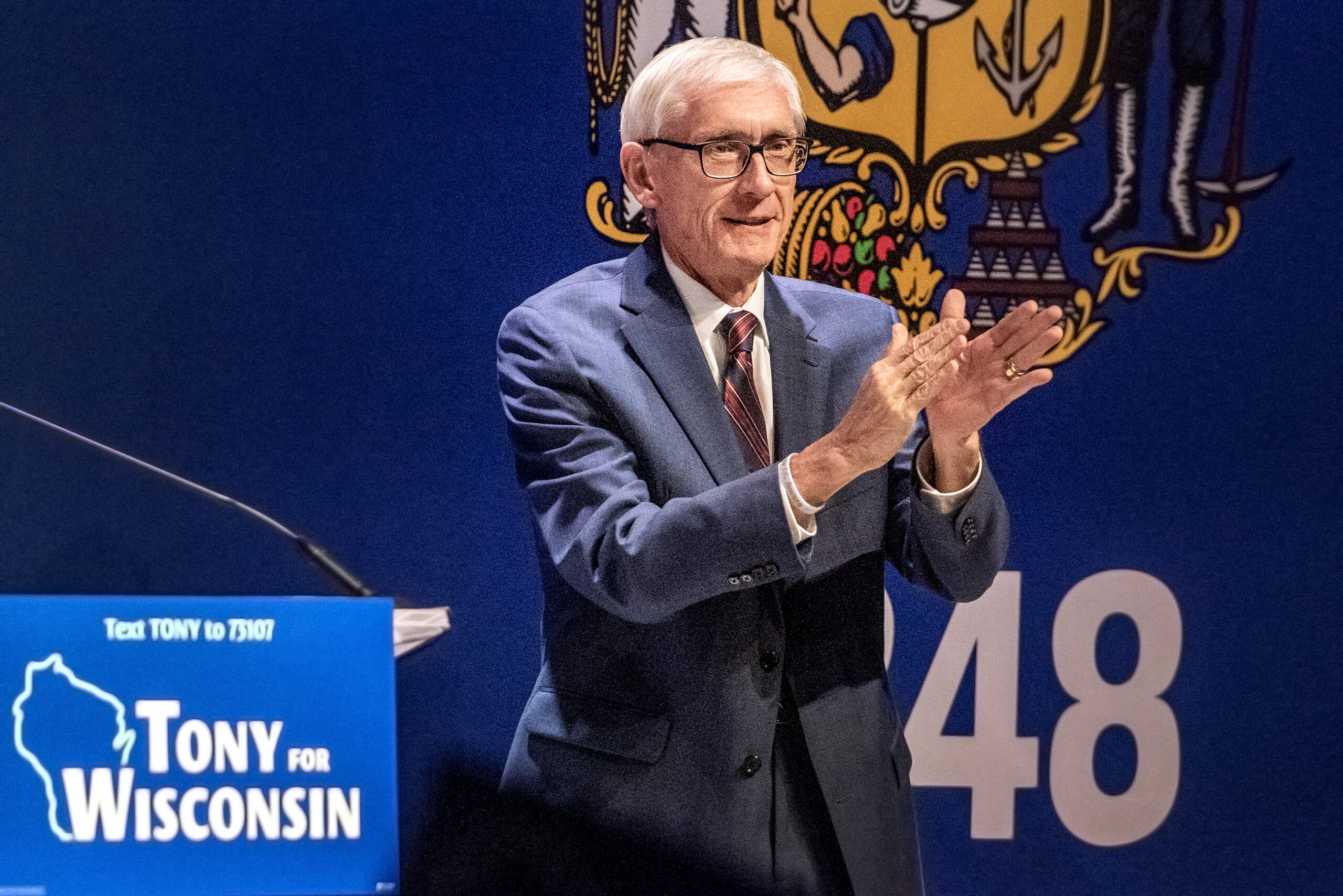 Tony Evers applauds while standing on a stage in front of a Wisconsin flag background.