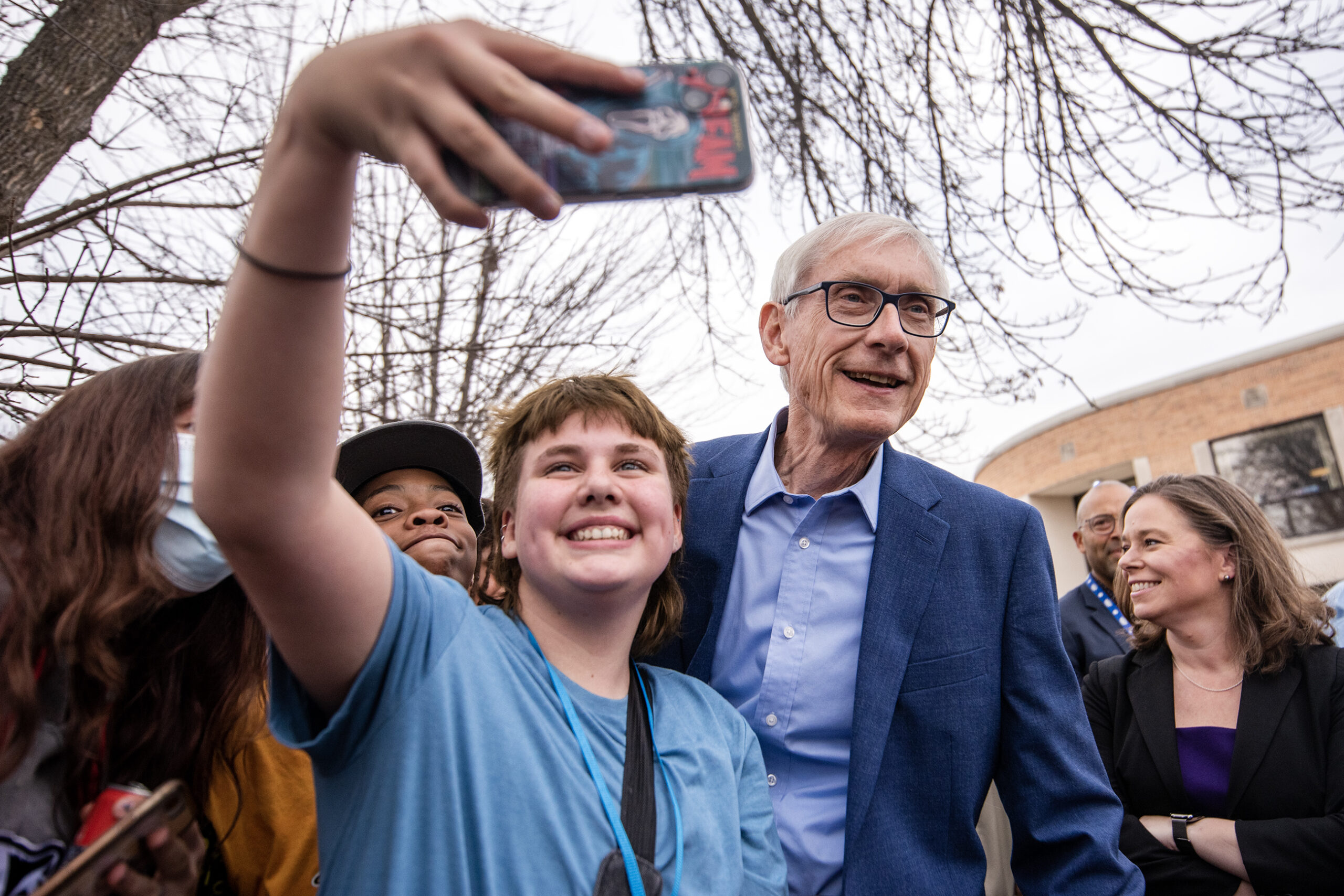 A student holds up a phone to take a selfie with Gov. Evers. Other students gather around to get in the photo.