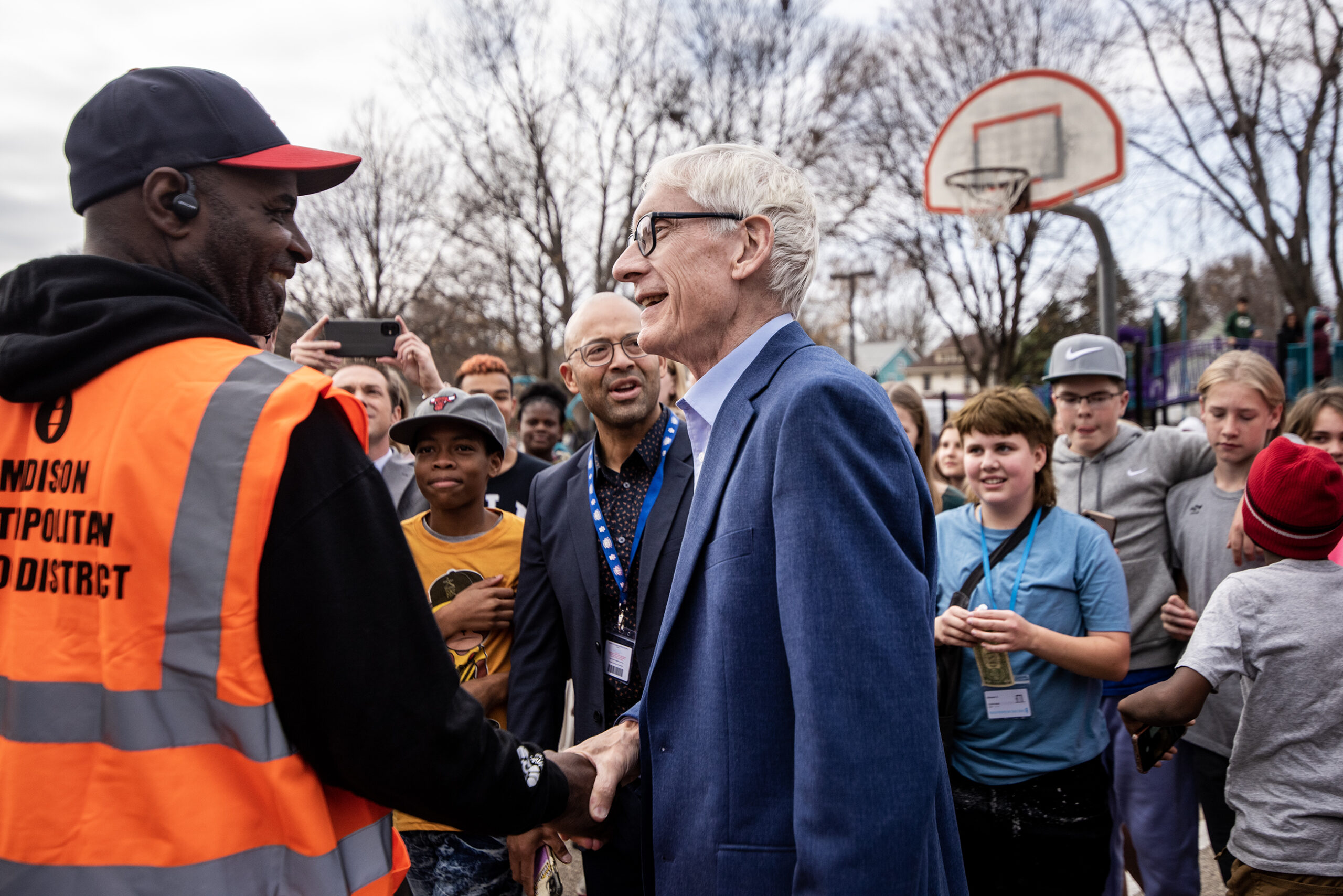 Gov. Evers shakes hands with a worker in an orange vest on a playground surrounded by middle school students. A basketball hoop can be seen in the background.