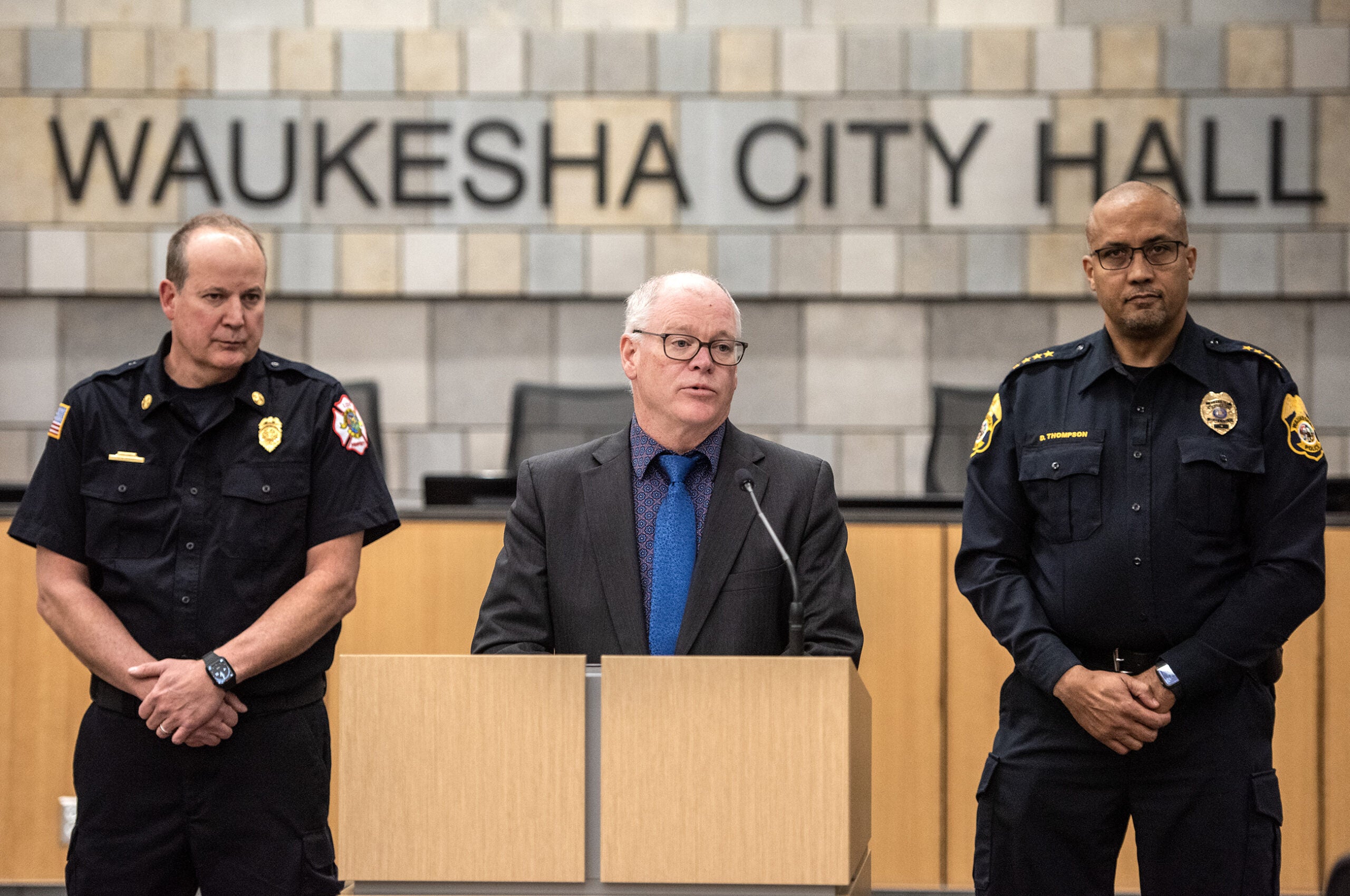Three men stand together. A sign says "Waukesha City Hall" behind them.